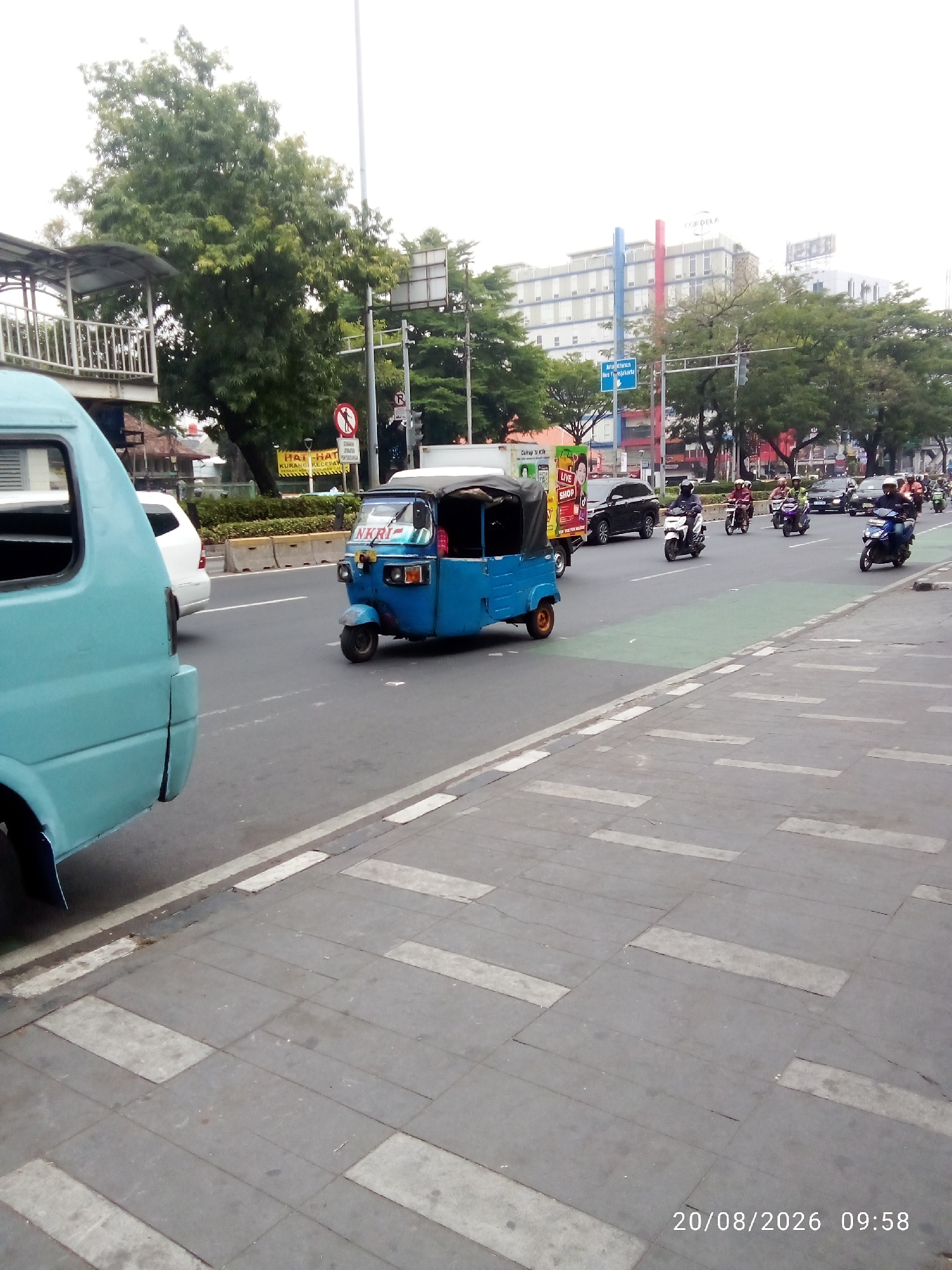 The image depicts a street scene in Central Jakarta, Indonesia, on August 20, 2026, at 09:58. A turquoise colored vehicle, possibly a van, is parked on the left side of the frame, partially obscuring the view. In the center of the road, a blue three-wheeled vehicle, commonly known as an "ojek" or "bajaj", is in motion. Several motorcycles, some with riders, are also traveling on the road, with some heading towards the background and others in the foreground. The road is paved and has distinct lane markings, including a green painted lane. On the right side of the road, there is a sidewalk with a patterned pavement. In the background, trees line the street, and beyond them, modern buildings can be seen, including one with the word "CORDELA" visible on its facade. Traffic signals and street signs are present, indicating a busy urban environment. The sky is overcast, contributing to a generally muted lighting condition.