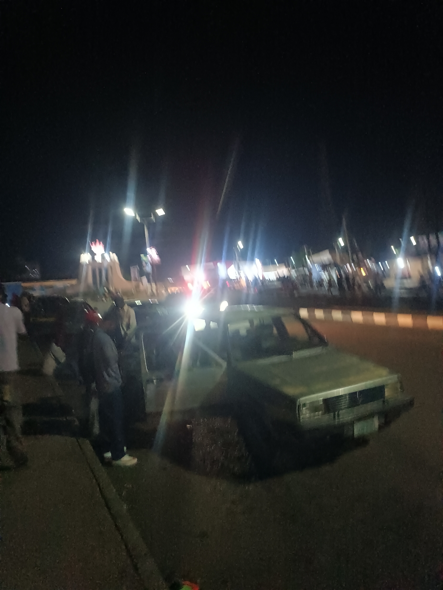 At night in Barakin Lerawi, Nigeria, an older model, light-colored car is parked on a paved road near a curb. A group of people stands near the car and on the curb. Streetlights illuminate the scene, and in the background, a structure is visible with what appears to be a crown at the top. There is a roadside barrier marked with reflective white and orange stripes. Pedestrians are visible farther in the background. The scene appears to be in a public area with moderate activity.