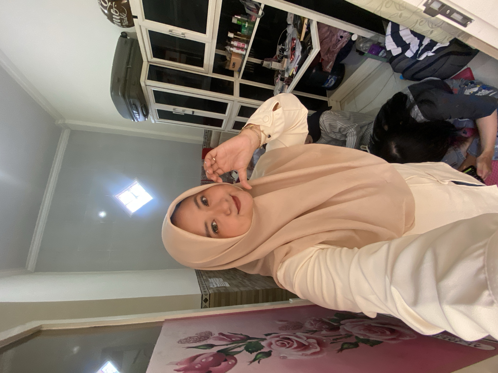 A woman wearing a light-beige hijab and white shirt is positioned in the foreground. Behind her, another person is partially visible, with dark hair, wearing a grey striped shirt and darker clothing. The setting appears to be an indoor space with a combination of furniture and storage, including a white cabinet with multiple compartments, some containing items. The backdrop features an off-white wall, a ceiling with recessed lighting, and a portion of a flower-patterned surface. This appears to be a residential space. There is no information to specify the city of Jember, Indonesia.