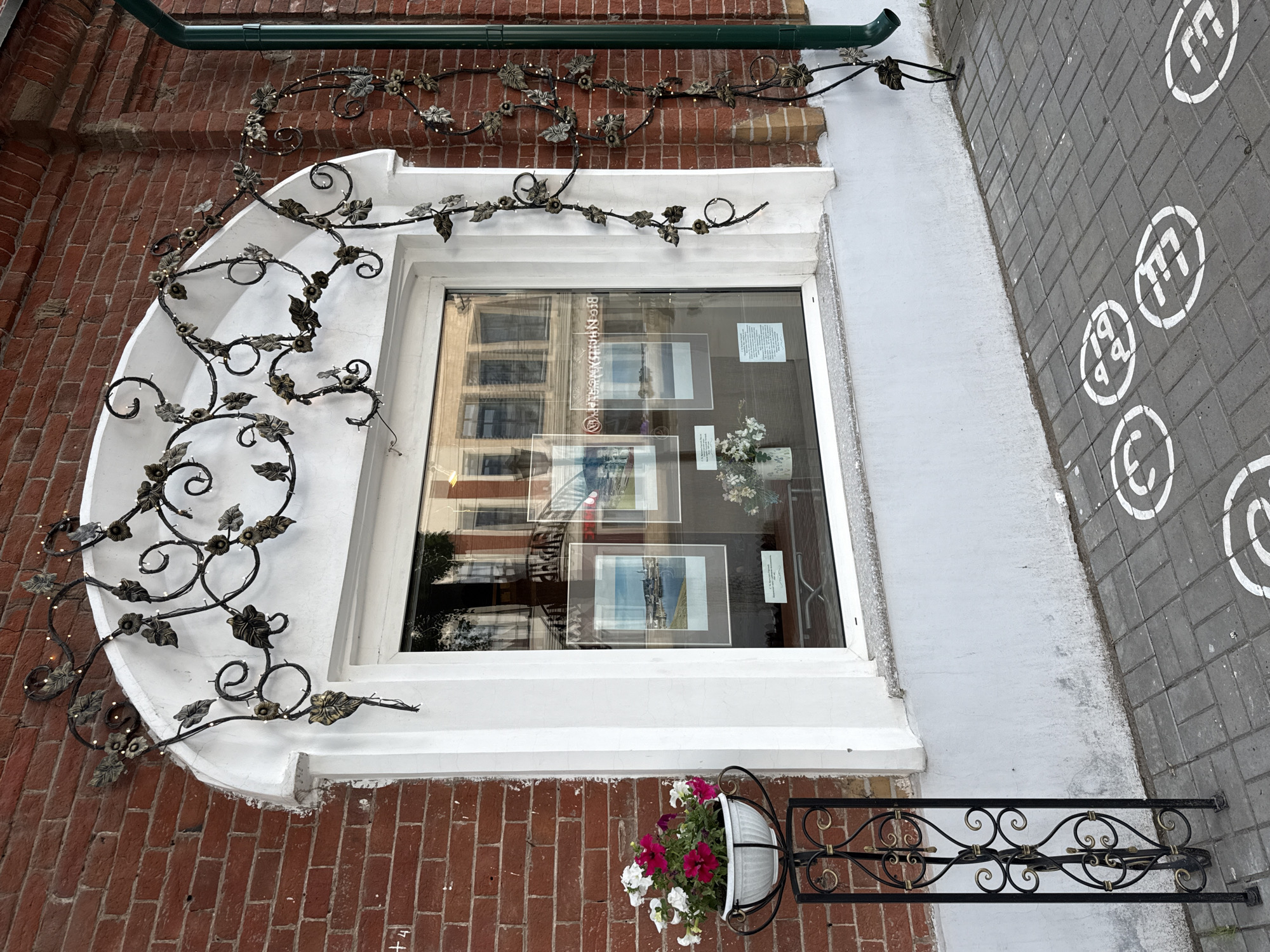 The image shows the exterior of a building with a red brick facade and a large arched window. Above the window, there is a black wrought iron decorative element with leaf and flower motifs, illuminated by small fairy lights. A green downspout is visible on the right side of the building.
To the left of the window, a black metal stand holds a white planter filled with pink and white flowers. The window itself displays several framed pictures, suggesting it might be a shop or gallery. The reflection in the window shows buildings across the street.
The ground in front of the building is paved with grey interlocking bricks, and several white circular markings with numbers or symbols are painted on the pavement, possibly indicating parking spaces or a designated area. The overall scene is set in an urban environment.
This scene is located in Votkinsk, Russia. Specifically, it is the facade of the Museum of History and Local Lore of Votkinsk, also known as the "House of the Composer Tchaikovsky's Mother." The building is located at 57 K. Marx Street.