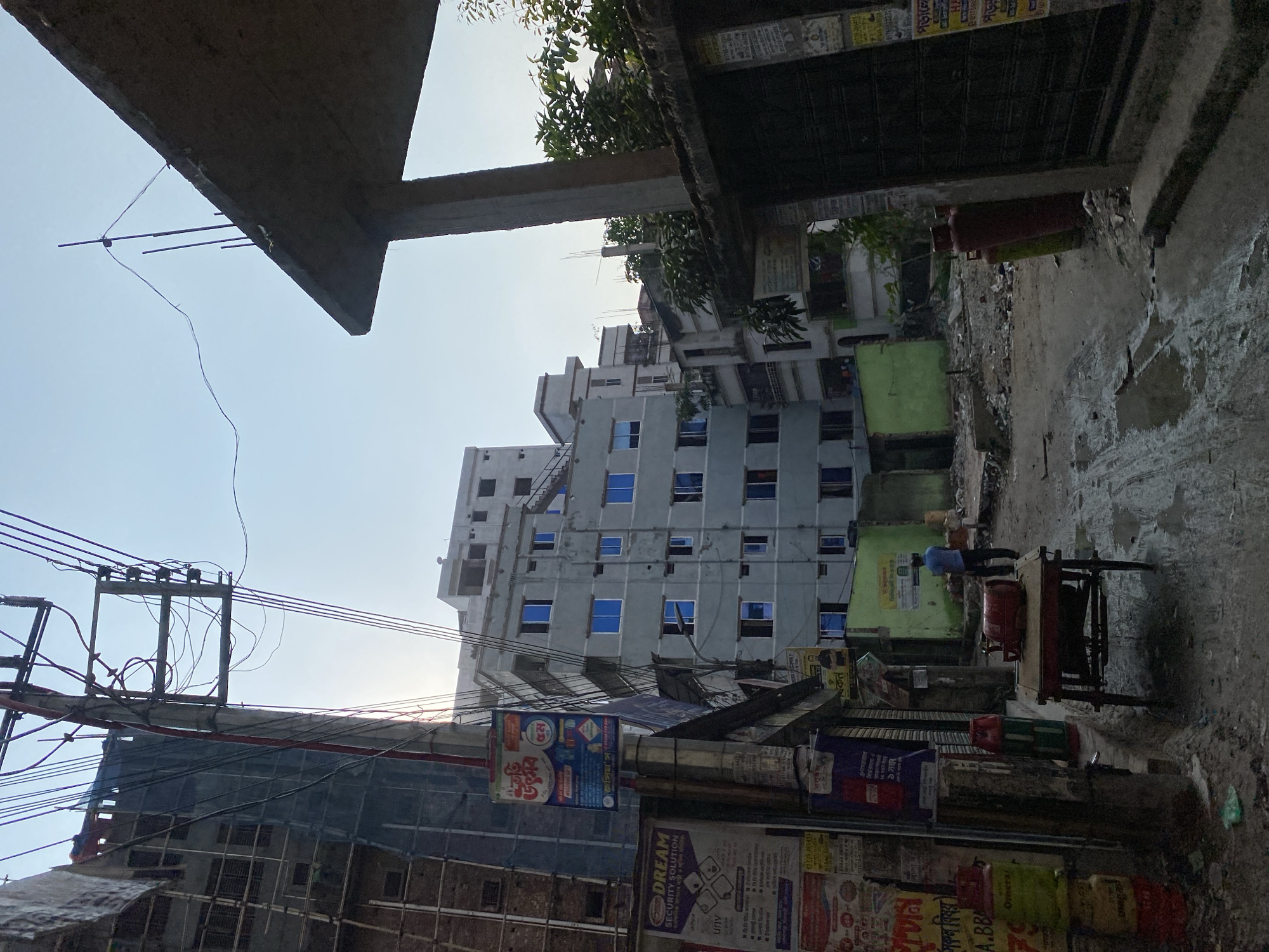 The image captures a scene in Dhaka, Bangladesh. In the foreground, there are poles with electrical wires crisscrossing, along with advertisements and posters attached to buildings. Construction is evident with scaffolding and unfinished buildings. A taller building with blue-tinted windows rises in the background. A man is seen pushing a cart carrying propane tanks on a narrow street. The environment is urban, with signs of disrepair and development.