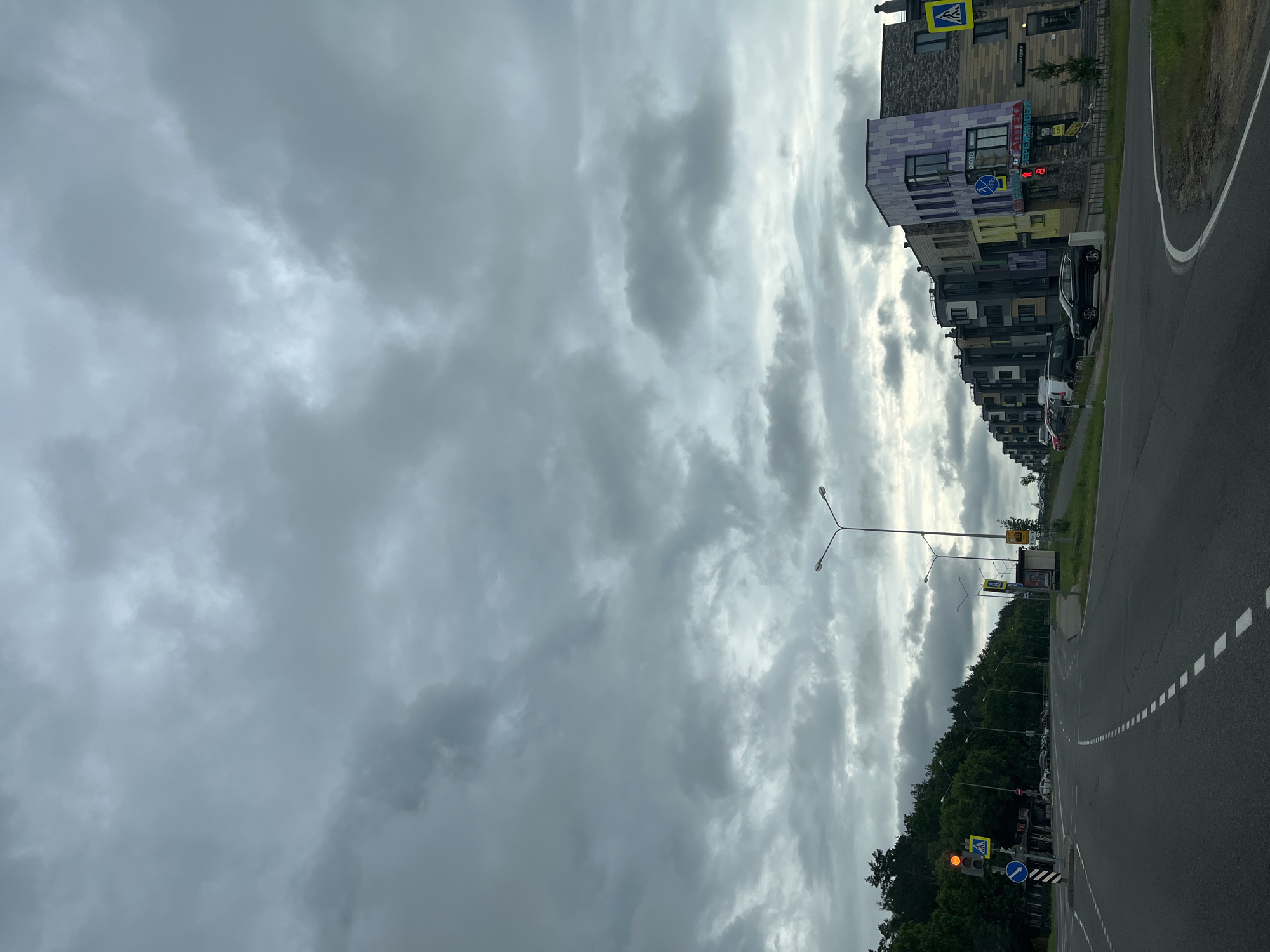 The image depicts a cloudy day in a suburban area. A paved road curves to the right, with white dashed lines marking the lanes. On the left side of the road, a traffic light shows a yellow signal, and a pedestrian crossing sign is visible. Further down, a densely wooded hill rises. Opposite the hill, on the right side of the road, is a row of modern multi-story buildings with varied architectural styles and colorful facades. Cars are parked along the buildings, and there are signs, including one for a pharmacy and another with a bicycle icon. Streetlights are positioned along the road. The sky is filled with thick, grey clouds, suggesting overcast weather.