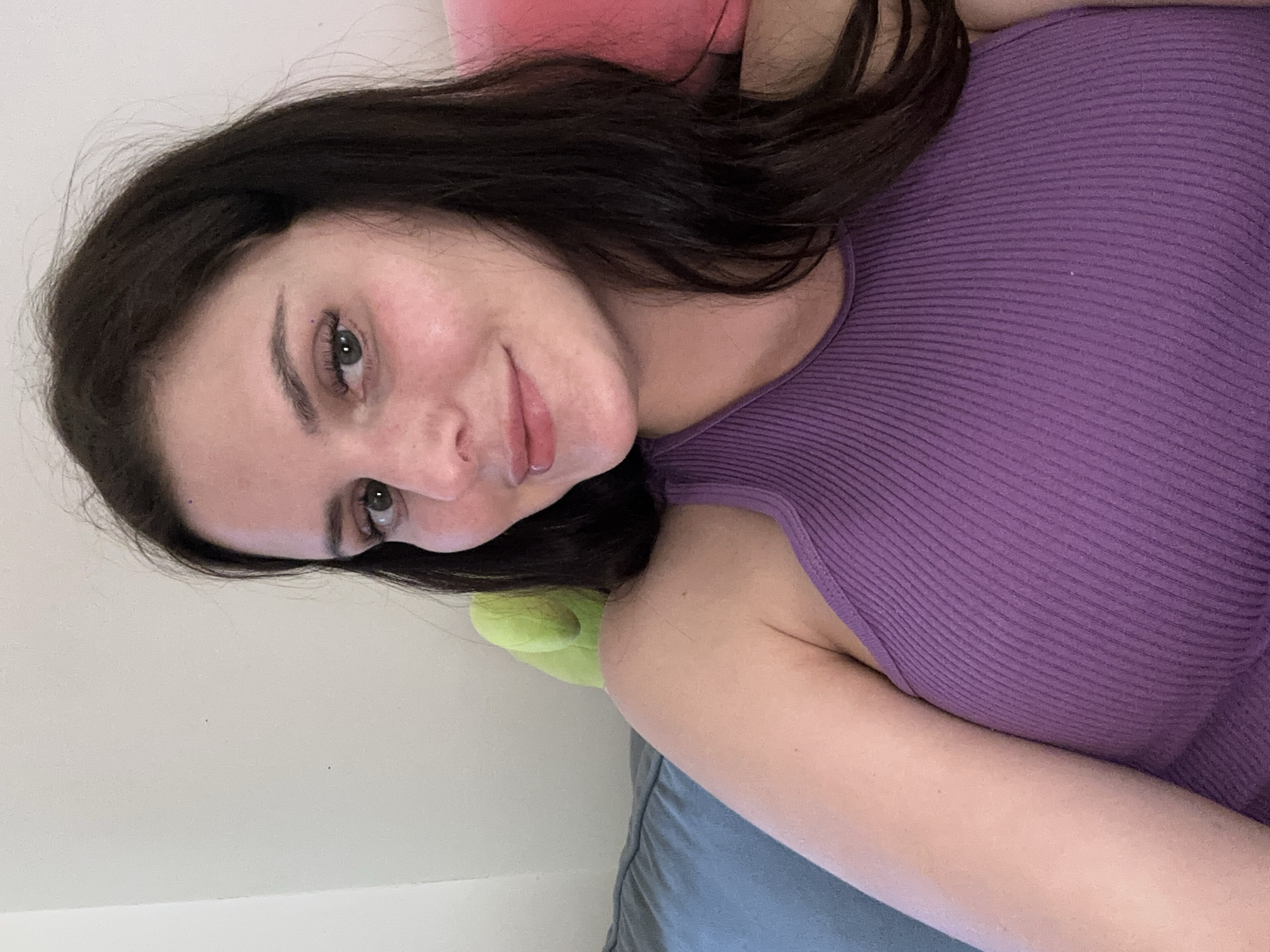A young woman with dark brown hair and green eyes looks at the camera with a slight smile. She is wearing a purple ribbed tank top. Behind her, a blue couch is visible, along with a portion of a bright pink pillow and a green stuffed animal. The background is a plain, light-colored wall. The image appears to be a selfie taken indoors.