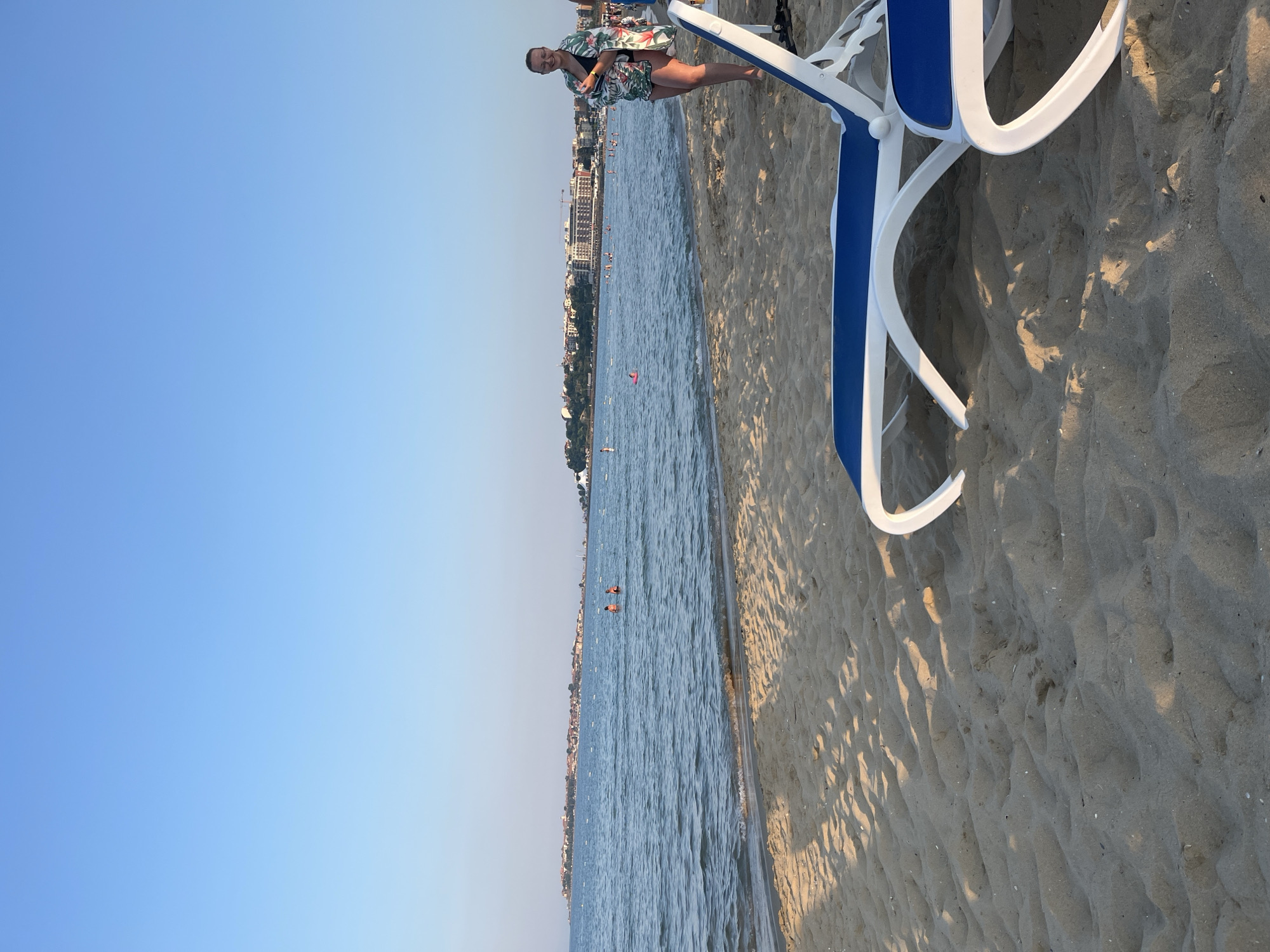 The image depicts a scenic view of the Nesebar, Bulgaria coastline. The foreground features a sandy beach with two blue and white lounge chairs, partially visible. The sea, with gentle waves, stretches towards the horizon. In the distance, the historic town of Nesebar is visible, characterized by its cluster of buildings and churches situated on a peninsula. Several people are scattered across the beach and in the water, some swimming and others relaxing. A woman wearing a floral sarong over a swimsuit stands on the beach, facing the camera. The sky is clear and blue, indicating a sunny day. The overall scene conveys a tranquil beach atmosphere.
