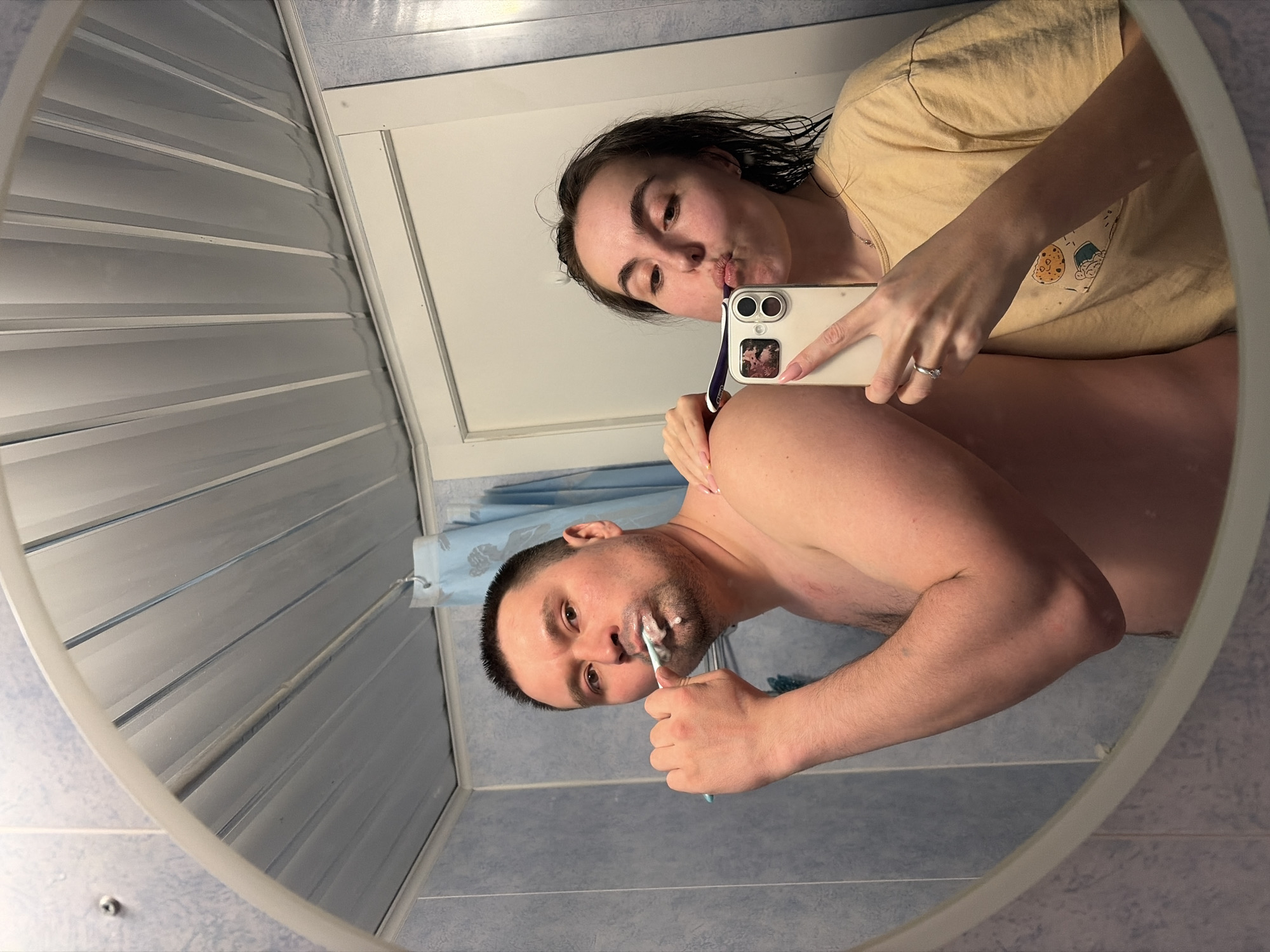 A selfie of a couple is reflected in a bathroom mirror. The man is shirtless and brushing his teeth. The woman is wearing a yellow t-shirt and holding a smartphone. The background consists of blue and white tiled walls. It is not possible to determine the location of the city or any other information regarding Izhevsk, Russia.