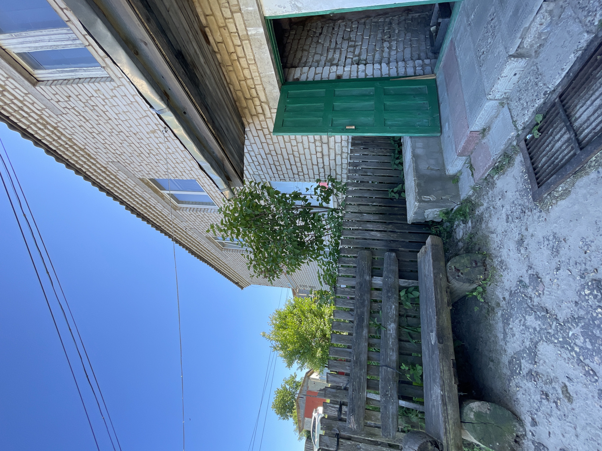 The image captures a sunlit outdoor scene in the village of Krasnye Baki, Russia. A weathered, rustic wooden bench sits in the foreground on a patch of dusty ground, with a wooden fence behind it. To the right, a light-colored brick building with a corrugated roof features an open, bright green door revealing a brick interior. A tree with lush green foliage grows beside the building. The sky above is a clear, vibrant blue.