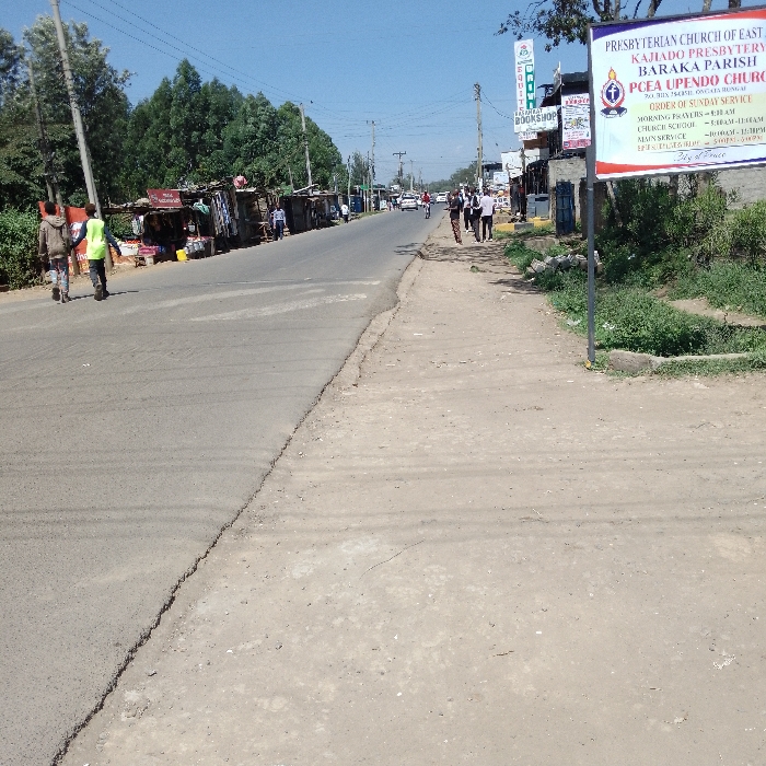 The image depicts a daytime street scene in Nkaimurunya ward, Kenya, under a clear blue sky. An asphalt road, exhibiting surface wear, extends from the foreground into the distance, appearing to curve slightly to the right. On the left side of the road, a continuous line of roadside vendor stalls, constructed from corrugated metal and wood, is present. Behind these stalls are dense green trees and utility poles with overhead power lines. Two individuals are observed walking on the asphalt road on the left, moving away from the viewer; one wears a bright yellow-green reflective vest. To the right of the asphalt road is a wide, unpaved dirt shoulder with patches of green vegetation. A large white billboard sign, bearing text for the "PRESBYTERIAN CHURCH OF EAST KAJIADO PRESBYTERY BARAKA PARISH PCEA UPENDO CHURCH," is prominently positioned on this right shoulder. Further along the right side, a group of approximately five to seven pedestrians is walking on the dirt shoulder, moving away from the foreground. Behind them and further down the road, more structured commercial buildings are visible, one with a sign indicating "BOOKSHOP." In the mid-ground and background, several vehicles are present on the road, and additional utility poles line the street.