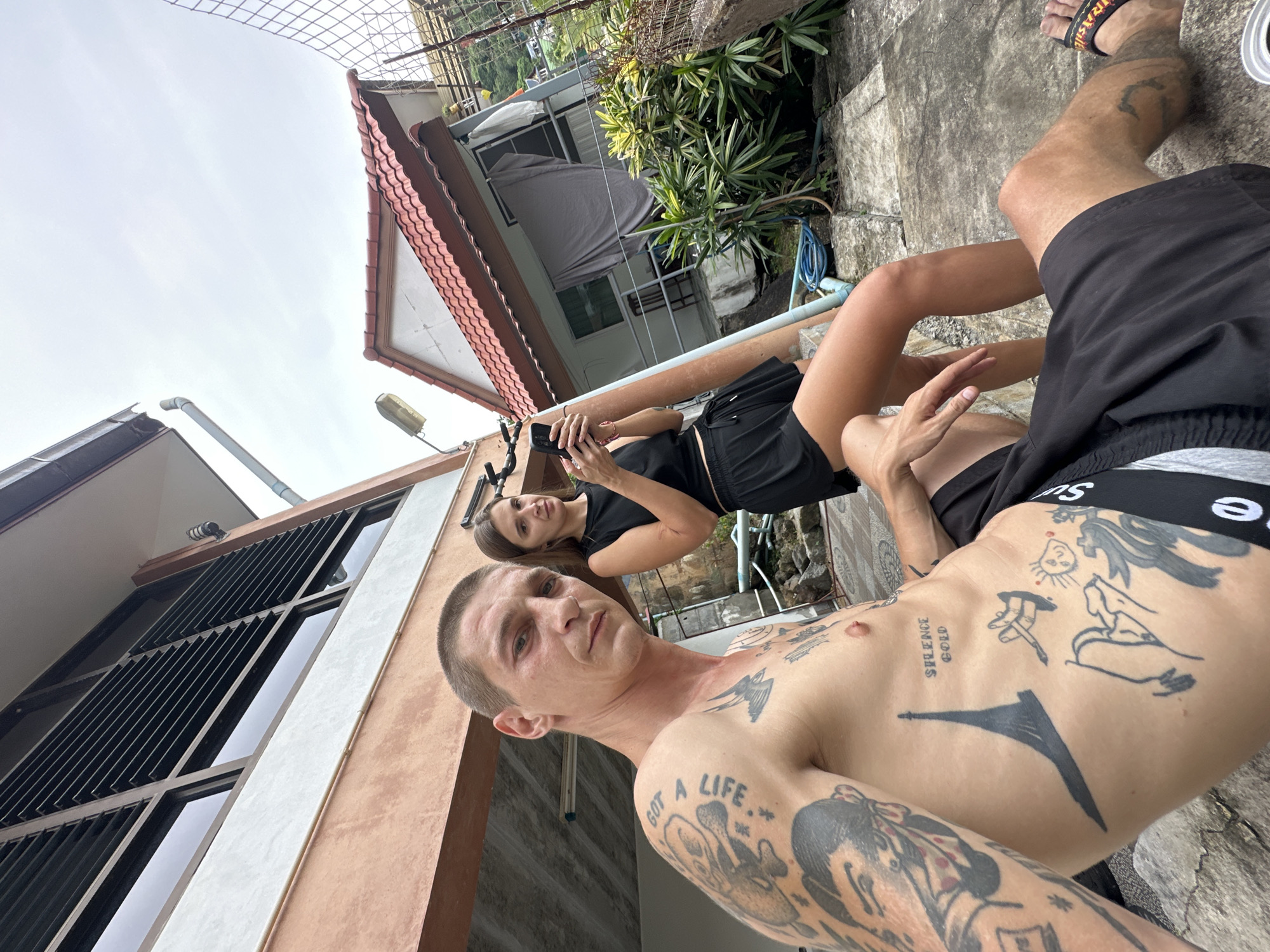 Two individuals, a man and a woman, are pictured in an outdoor setting. The man, with numerous tattoos on his torso and arm, is in the foreground, looking towards the camera. He is shirtless and wearing black shorts. The woman stands behind him, holding a smartphone and looking down at it. She is wearing a black crop top and black shorts. The background consists of a building with a distinctive tiled roof and louvred windows, and lush green vegetation. The ground is paved with concrete. The scene appears to be in a residential area. The overall atmosphere is casual.