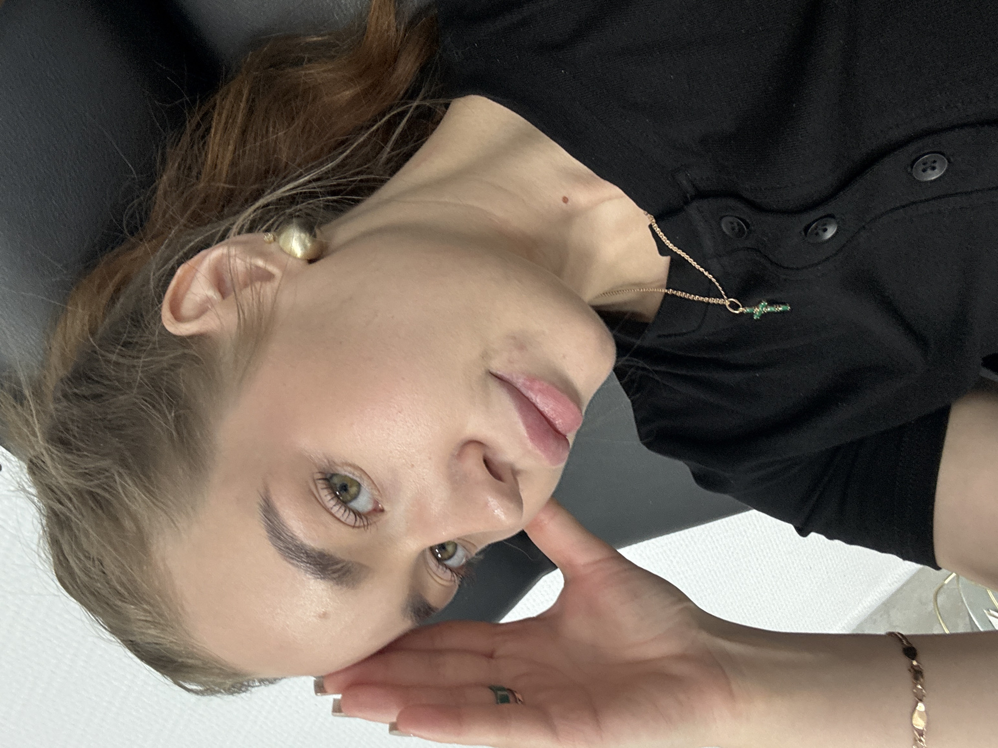 A woman is lying down. She has fair skin, green eyes, and light brown hair with some auburn highlights. She is wearing a black button-down shirt, a gold necklace with a green cross pendant, and gold earrings. Her left hand is near her face, and she wears a ring on her ring finger and a bracelet on her wrist. The background appears to be an examination table, with a white surface visible beneath her. The location is not determined from this image.