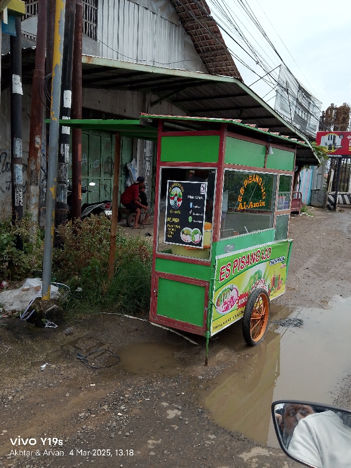 The image shows a green food cart parked in front of a building in Jatibarang, Indonesia, at coordinates -6.468 latitude, 108.296 longitude. The cart is selling "Es Pisang," which translates to banana ice cream. The cart has a sign with an image of banana ice cream and a price of "Rp. 8000." There is a man in a blue shirt standing near the cart, and another man in a red shirt is out of focus in the background. The ground is muddy and there is a puddle of water in front of the cart. The scene is a street in a residential area. The image was taken on a sunny day. There is a power line running across the top of the image.