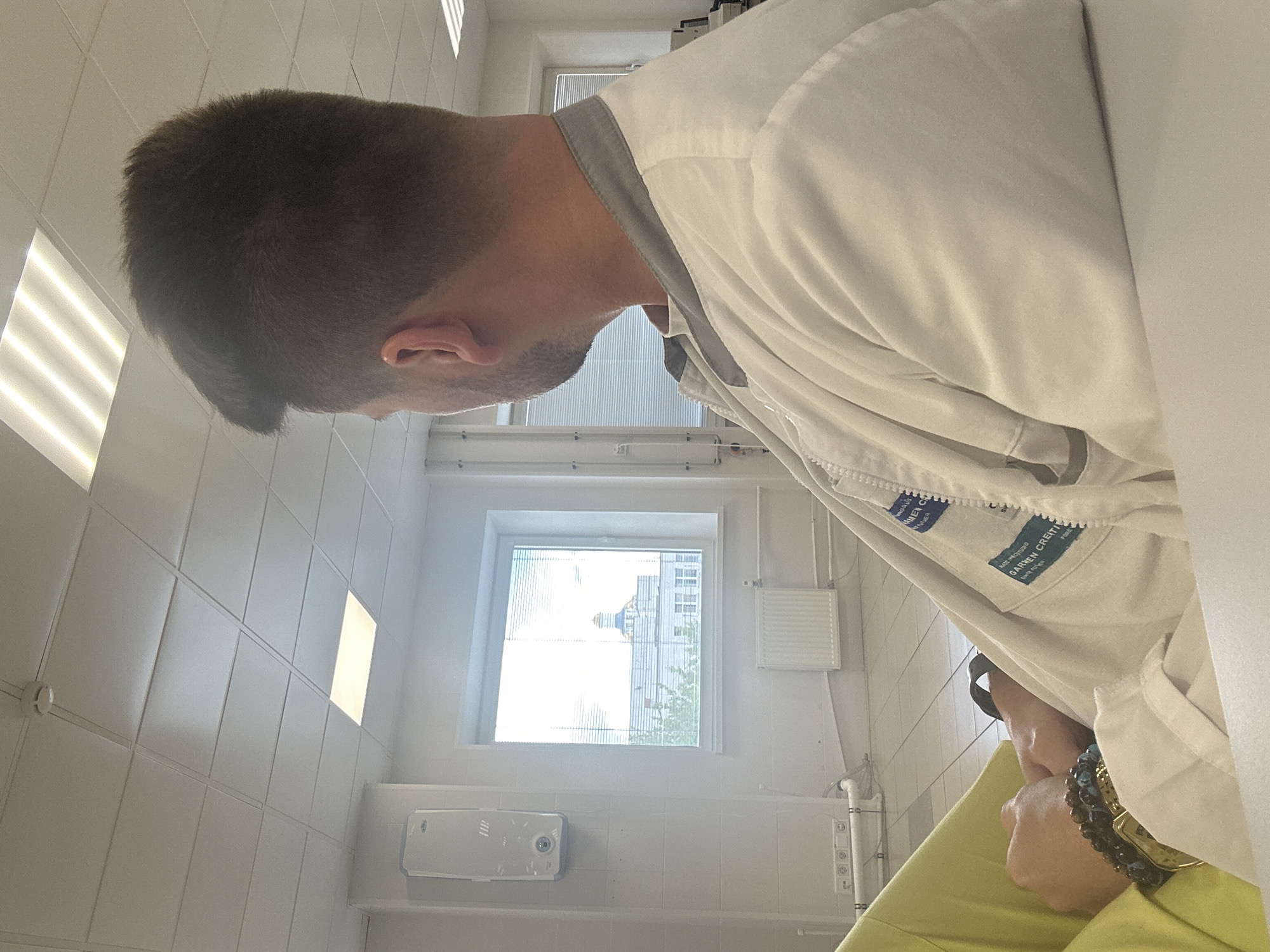 A man with short, dark hair is wearing a white lab coat with a grey collar. A tag with the name "GARNEN CREITI" is visible on the coat. The man is looking down and to his left. The scene appears to be inside a medical facility or laboratory, as indicated by the tiled walls, a window with blinds, medical equipment, and the lab coat. Outside the window, a building with a dome-shaped roof is visible, suggesting a city landscape. Based on the architectural style of the domed building, it might be located in Moscow, Russia.