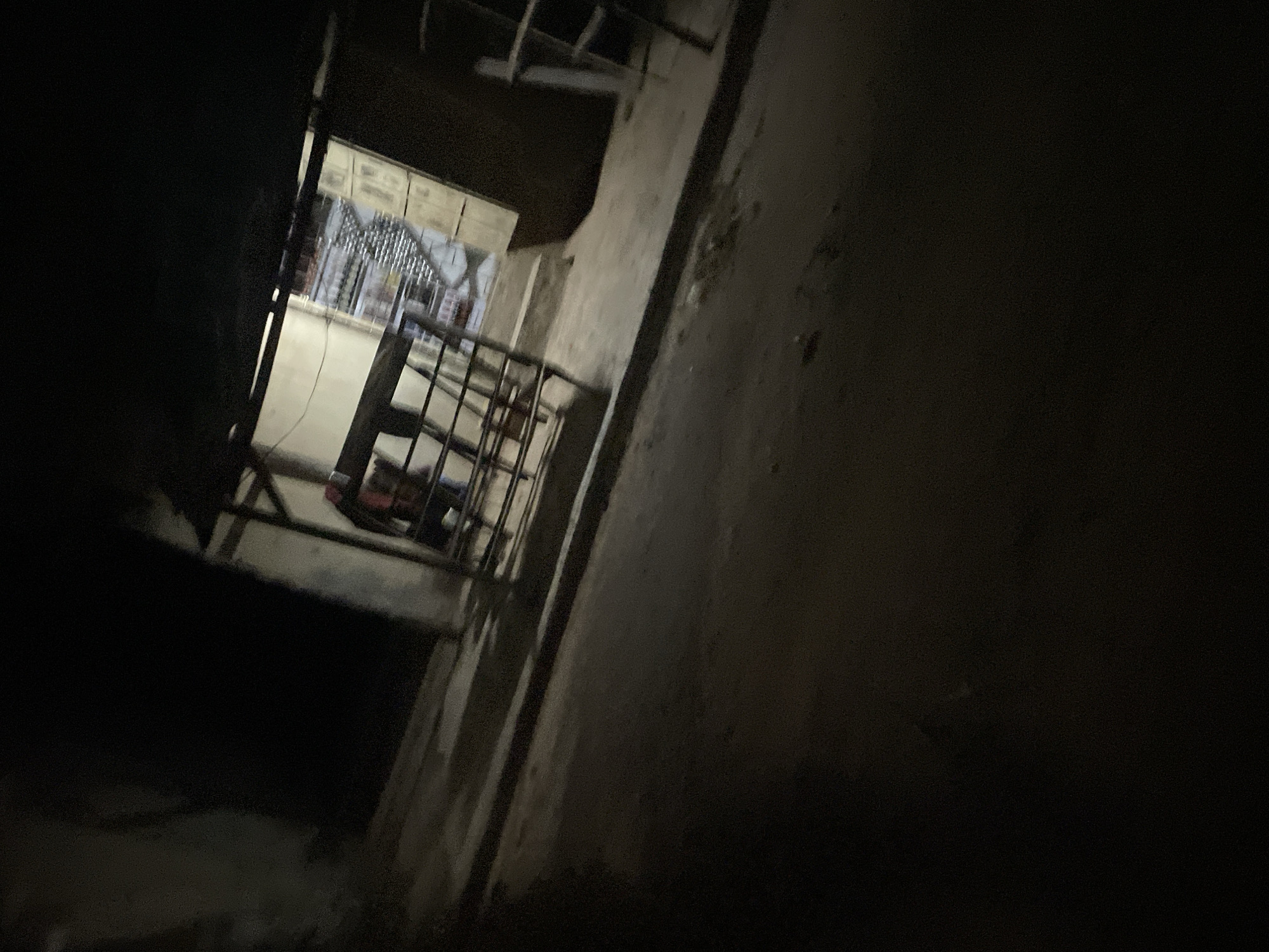 The image depicts a dimly lit scene, likely an outdoor market stall or a small shop at night in Lokoja, Nigeria. A metal grate or fence is in the foreground, separating the viewer from the interior of the stall. Inside the stall, shelves are visible, stocked with rows of bottled drinks. A person, partially obscured, appears to be behind the counter. The ground in front of the stall is paved with a rough surface. The overall lighting is low, with the interior of the stall being the brightest area.