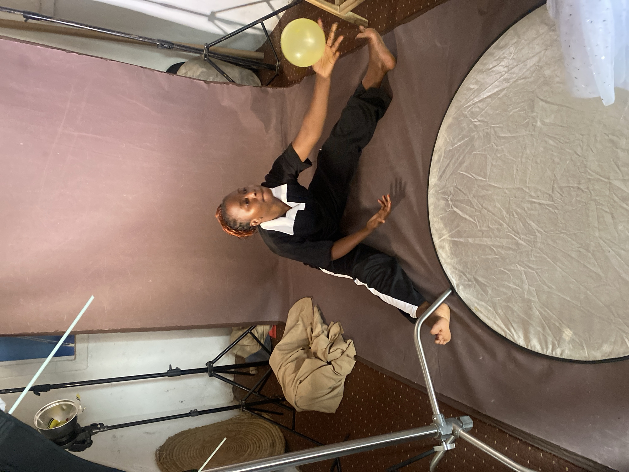 A full-length shot depicts a young person juggling a yellow balloon in what appears to be a studio or indoor photography setting. The individual, facing towards the camera, is dressed in black pants with a white stripe down the side and a black top with a white collar. They are barefoot. The studio has a backdrop of plum color on the left, with lighting equipment and various stands visible. A round reflector is placed on the right. Based on the person's physical features, they could be from Vom, Nigeria. The floor has a patterned brown carpet. The environment suggests a professional or semi-professional photographic setup.