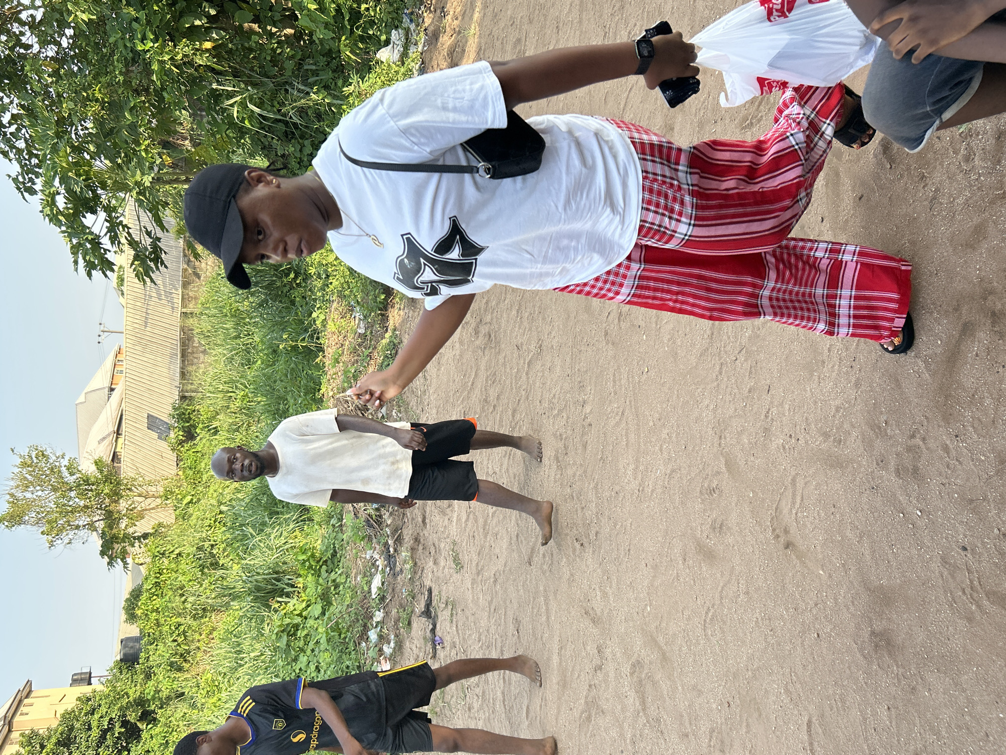 A woman wearing a black baseball cap, a white t-shirt with a black graphic design, red and white plaid pants, and black sandals is walking on a dirt path. She is carrying a white plastic bag in her right hand. She also has a black purse on her left shoulder and a black wristwatch on her left wrist. A man wearing a white t-shirt and black shorts is standing barefoot on the dirt path, looking toward the camera. A younger male wearing a black and yellow t-shirt and black shorts is also walking barefoot on the path, partially obscured at the bottom of the frame. The location is Ihiagwa, Nigeria, as the image's EXIF data includes the city name. The surrounding environment includes green vegetation, a building, and a clear sky.