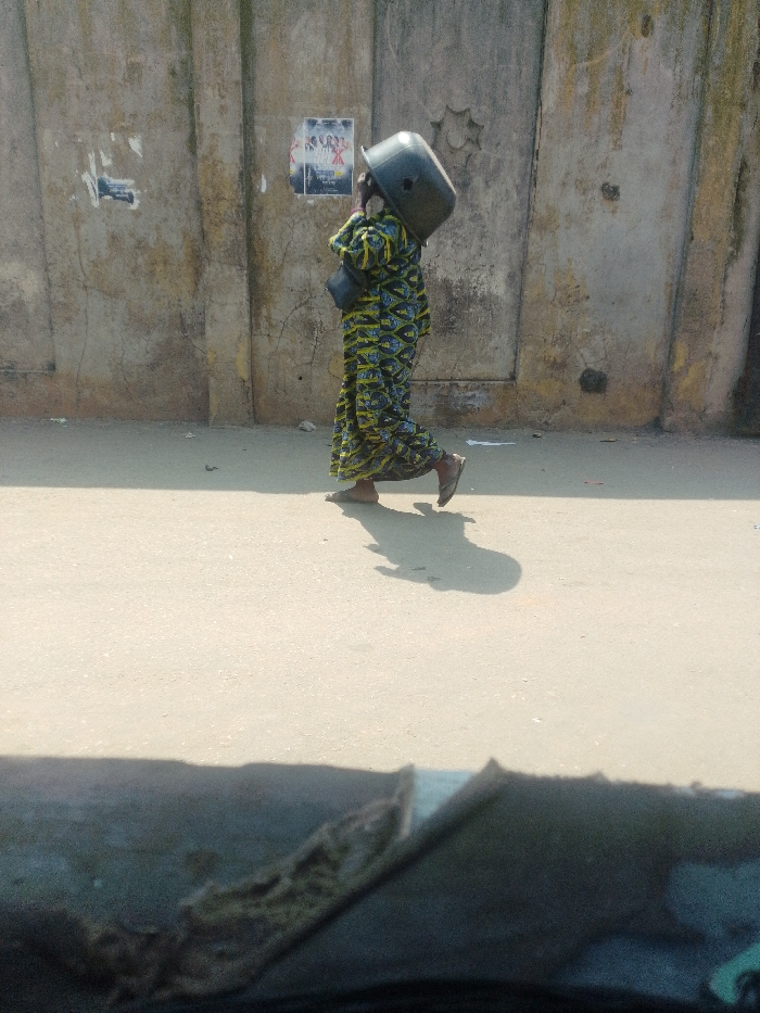 The image depicts an individual walking on a paved surface alongside a textured concrete wall under bright daylight in Agani, Nigeria.
**People:** One person is visible, walking from left to right across the frame. The individual wears a long, patterned garment in green, yellow, and black, and open-toed sandals. A dark strap, possibly from a shoulder bag, is visible over their left shoulder. Their shadow is cast prominently on the ground behind and to their right.
**Objects:** A large, dark grey or black plastic bucket is balanced on the individual's head. Two posters are affixed to the concrete wall; the left one is partially torn, and the right one, more intact, displays figures. A decorative, star-shaped architectural element is embedded in the wall to the right of the posters. The immediate foreground shows a blurred dark surface, possibly the interior of a vehicle.
**Actions:** The person is in motion, striding forward with the left foot planted and the right foot lifted mid-step. They are transporting the bucket on their head.
**Environment:** The scene is outdoors on a sunny day, indicated by clear shadows. The ground is a light-colored, even paved surface. The background consists of a tall, light brown or grey concrete wall, showing signs of wear, discoloration, and cracks.