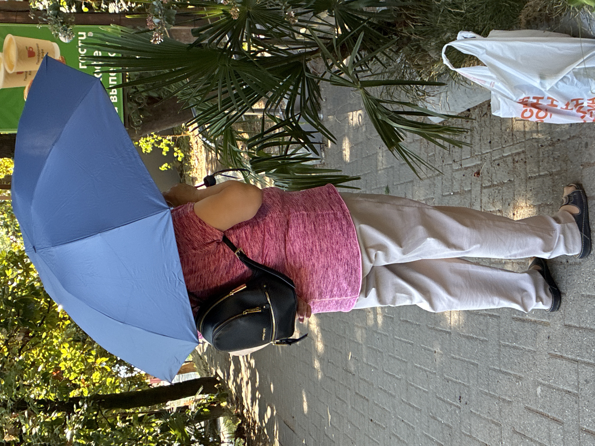 A person stands on a paved walkway under a large, blue umbrella. The person is wearing a pink, short-sleeved shirt, beige pants, and dark slip-on shoes. A small black backpack with gold hardware is worn on their back. To the right of the person, a palm frond plant is visible, and a white plastic shopping bag with red lettering is on the ground. In the background, trees with dappled sunlight and a green sign with Cyrillic text and images of coffee cups are present. The scene appears to be in an outdoor setting, possibly a park or a street cafe in Sochi, Russia, given the presence of palm trees and the Cyrillic signage.