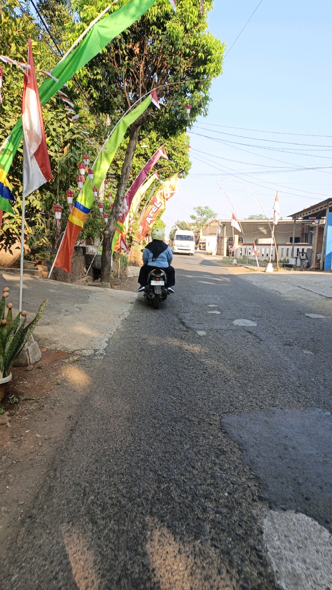 A person is riding a motorcycle down a road in Kuwasen, Indonesia. The person is wearing a blue jacket, black pants, and a yellow helmet. They are riding a black motorcycle with a license plate that reads "2379 JBH". The road is paved with asphalt and has some potholes. There are trees on the left side of the road with Indonesian flags hanging from them. There are houses and a white van on the right side of the road. The sky is blue and clear. The image appears to have been taken in the morning or afternoon, as there are shadows on the road.
