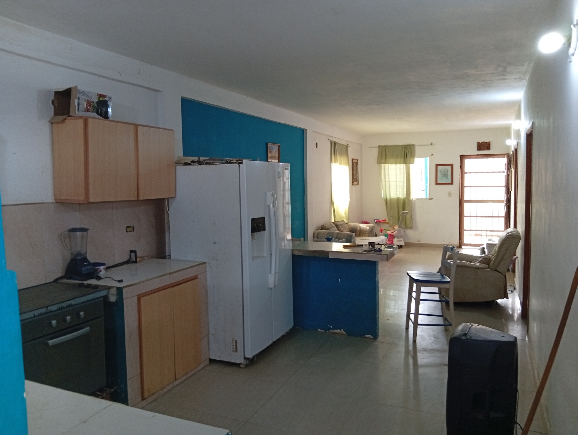 The image depicts the interior of a home or apartment in Juan Griego, Venezuela. The scene is an open-plan living area, with a kitchen transitioning into a living room and entryway.
The kitchen features light wood cabinetry, a gray stove, a white refrigerator, and a blue breakfast bar. The living room contains a sofa, armchair, and dining furniture. The entryway has a security screen door.
The environment appears modest. There are no people visible in the frame.