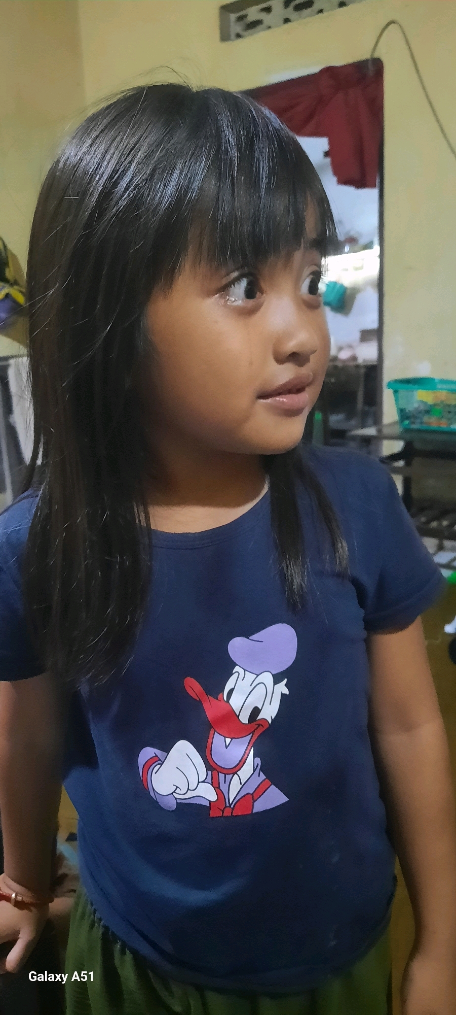A young Indonesian girl with dark hair and bangs, wearing a navy blue t-shirt with a cartoon duck character, stands in what appears to be a residential setting. The background is a pale yellow wall with a red curtain hanging behind her. A window with a decorative pattern is visible on the upper right. A green plastic basket is also in the background. The girl's expression is directed to her right. The lower left corner of the image displays "Galaxy A51", indicating the device used for the photograph. The location is identified as Jatitengah, Indonesia.