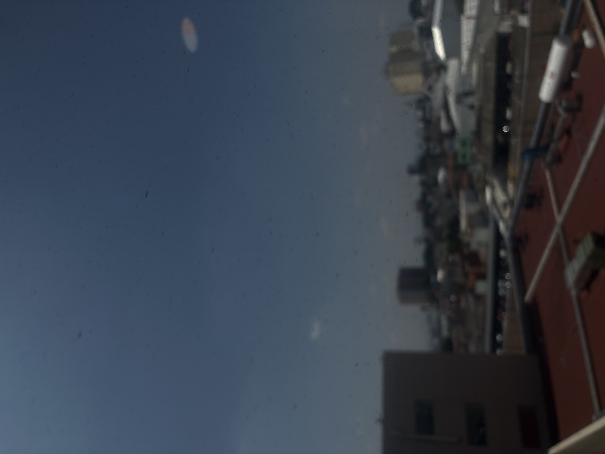 A rotated photograph, taken through a dirty window, captures a blurry cityscape under a blue-grey sky during the daytime. The image is oriented 90 degrees clockwise from its natural perspective, with the sky dominating the upper left and central portions when viewed correctly. Numerous small, dark specks of dust or dirt are visible on the foreground glass surface. A prominent bright, elongated, orange-white light flare, likely a reflection, appears in the upper-left of the sky.
The lower-right section (when correctly oriented) features an out-of-focus urban landscape, showing indistinct buildings, some with reddish roofs and others as dark, angular structures. Scattered within this blurry cityscape are several small, circular, bokeh-like reflections. A white cylindrical object is visible in the cityscape, but any text or logo on it is unreadable due to blur. The scene depicts a static view from an elevated position, likely over a city, consistent with the provided context of Puebla, Mexico. There is no discernible activity taking place.