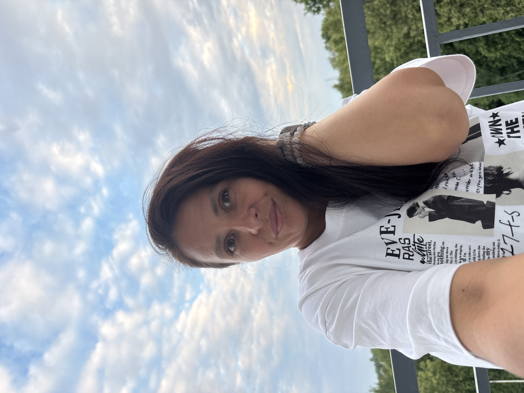 A woman takes a selfie outdoors with a cloudy sky in the background. She has long, dark hair and is wearing a white t-shirt with black text and an image. She is leaning on a railing, and green trees are visible behind her. The location is identified as Chaikovsky, Russia.