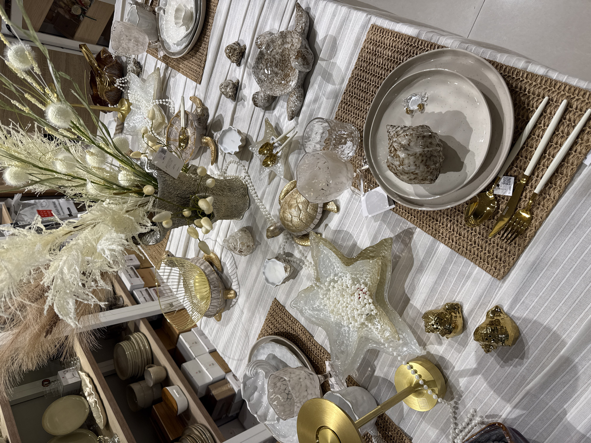 The image depicts a tabletop display, seemingly staged for a home decor or lifestyle advertisement. The scene is set against a background of vertically striped white fabric, likely a tablecloth. Several woven rectangular mats serve as placemats, adding a textural contrast.
The table is laden with decorative objects, many with a nautical or beachy theme. Ceramic turtles of varying sizes and colors are prominent, along with seashells and sea star-shaped bowls filled with white beads. A large floral arrangement with dried grasses and seed heads occupies a significant portion of the left side of the frame.
Place settings are arranged on some of the mats, including plates, flatware, and glassware. The colors are muted and neutral, with whites, creams, and golds dominating. Some price tags are visible on the items, suggesting they are for sale in a retail setting. A wooden shelving unit with additional tableware and decorative objects is visible on the left side of the image.
The scene appears to be indoors, possibly in a showroom or retail store. There is no visible presence of people. There is no forensic evidence to conclude that the location is in the city of Sochi, Russia, or that it is related to the city in any way.