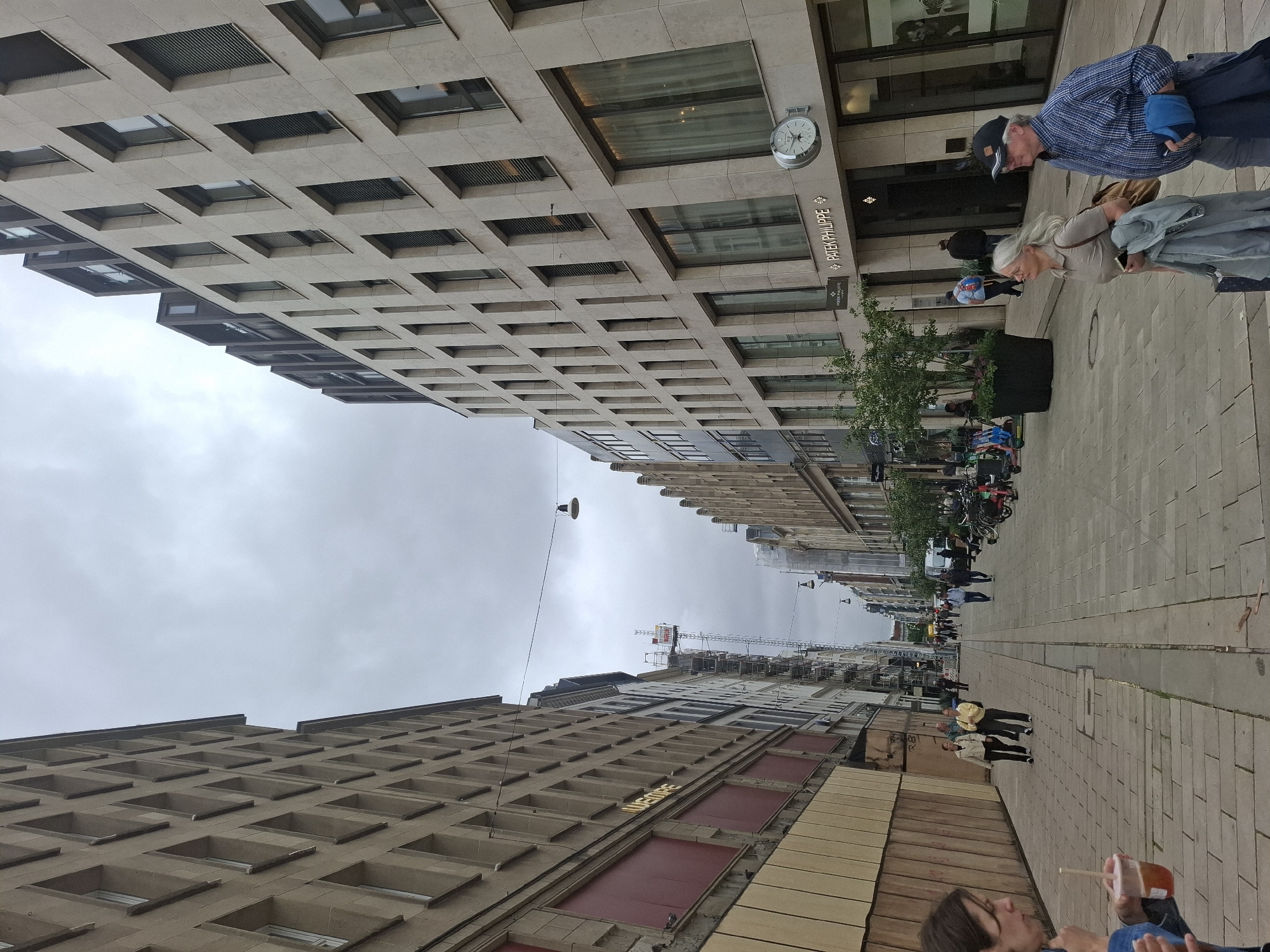 The image captures a street scene in Hamburg, Germany, likely on a cloudy day. The perspective is from street level, looking down a pedestrianized thoroughfare lined with modern buildings.
On the left, a building with a beige facade features rows of recessed windows and large, boarded-up sections with red material. The word "WEMPE" is visible on this building. The sidewalk in front of it is paved with large, rectangular stone slabs.
On the right, a contemporary building with a lighter beige stone facade rises several stories. It has numerous dark-framed windows and a prominent entrance with signage reading "PATEK PHILIPPE." A large round clock is mounted on the facade above the entrance. The sidewalk here is also paved with stone slabs.
Several people are visible walking along the street. In the foreground on the left, a person with dark hair is holding a drink. Further down the street, a couple walks together, and an older couple strolls towards the right side of the frame. Other pedestrians are scattered throughout the scene, some near parked bicycles.
In the background, the street continues, with more buildings and some trees in planters. A construction crane is visible in the distance, suggesting ongoing development or renovation in the area. Streetlights are suspended across the street. The overall atmosphere is one of urban activity on a overcast day.