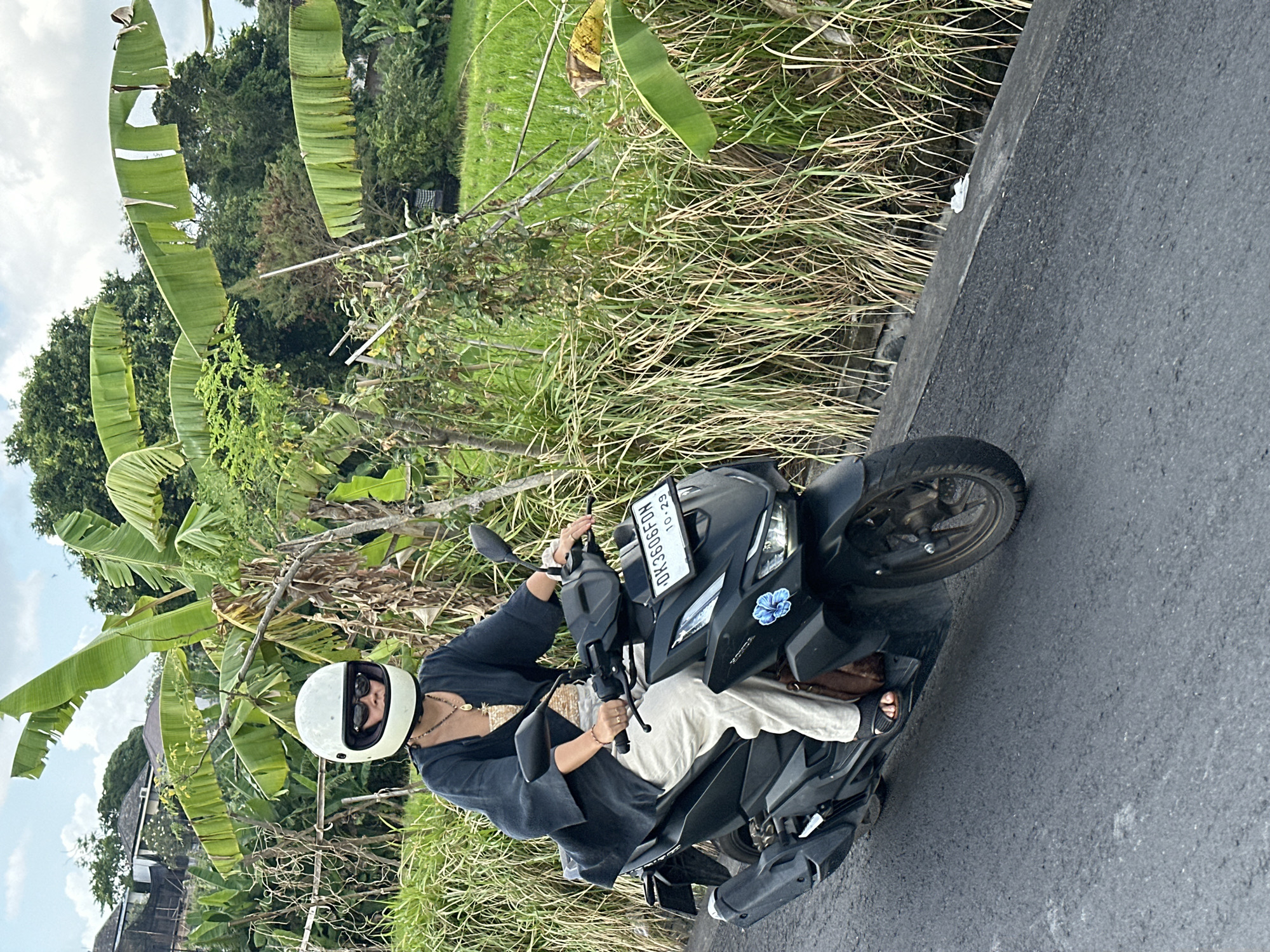 A person wearing a white helmet and sunglasses is riding a black scooter on a paved road. The person is wearing a dark, flowing top and light-colored pants, with a small brown bag slung over their shoulder. A blue flower sticker is visible on the front of the scooter. The road is bordered by tall, dry grass and lush green vegetation, including banana trees and what appears to be rice paddies, indicative of a rural or semi-rural setting in North Kuta, Indonesia. The sky is partly cloudy.