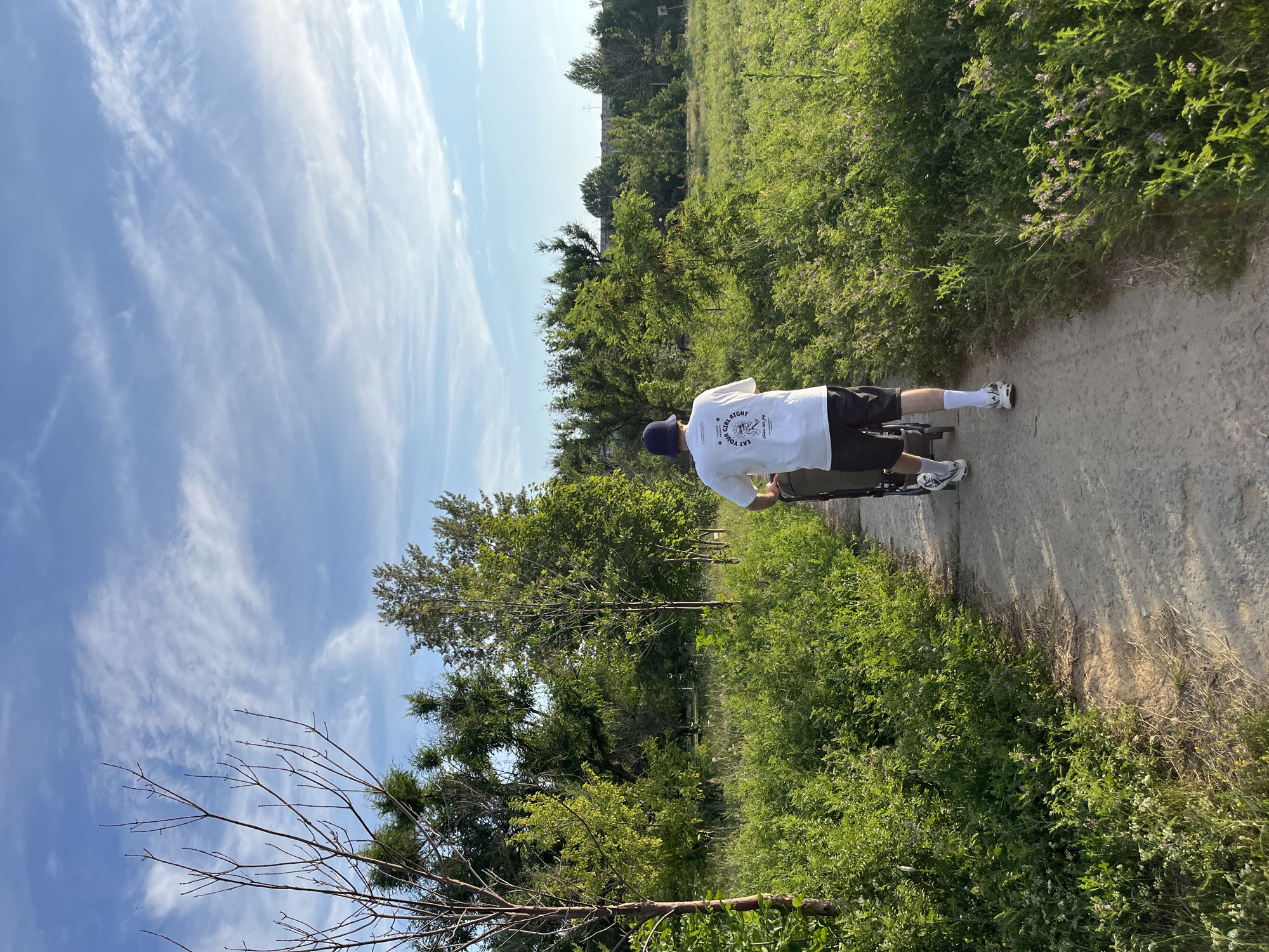 The image shows a person from behind, wearing a white t-shirt with text, dark shorts, white socks, and sneakers, pushing a stroller on a dirt path. The path is bordered by lush green vegetation and trees. The sky above is blue with scattered white clouds. The scene appears to be an outdoor recreational area. The location in the city of Kotovo, Russia is not discernible from the image alone.