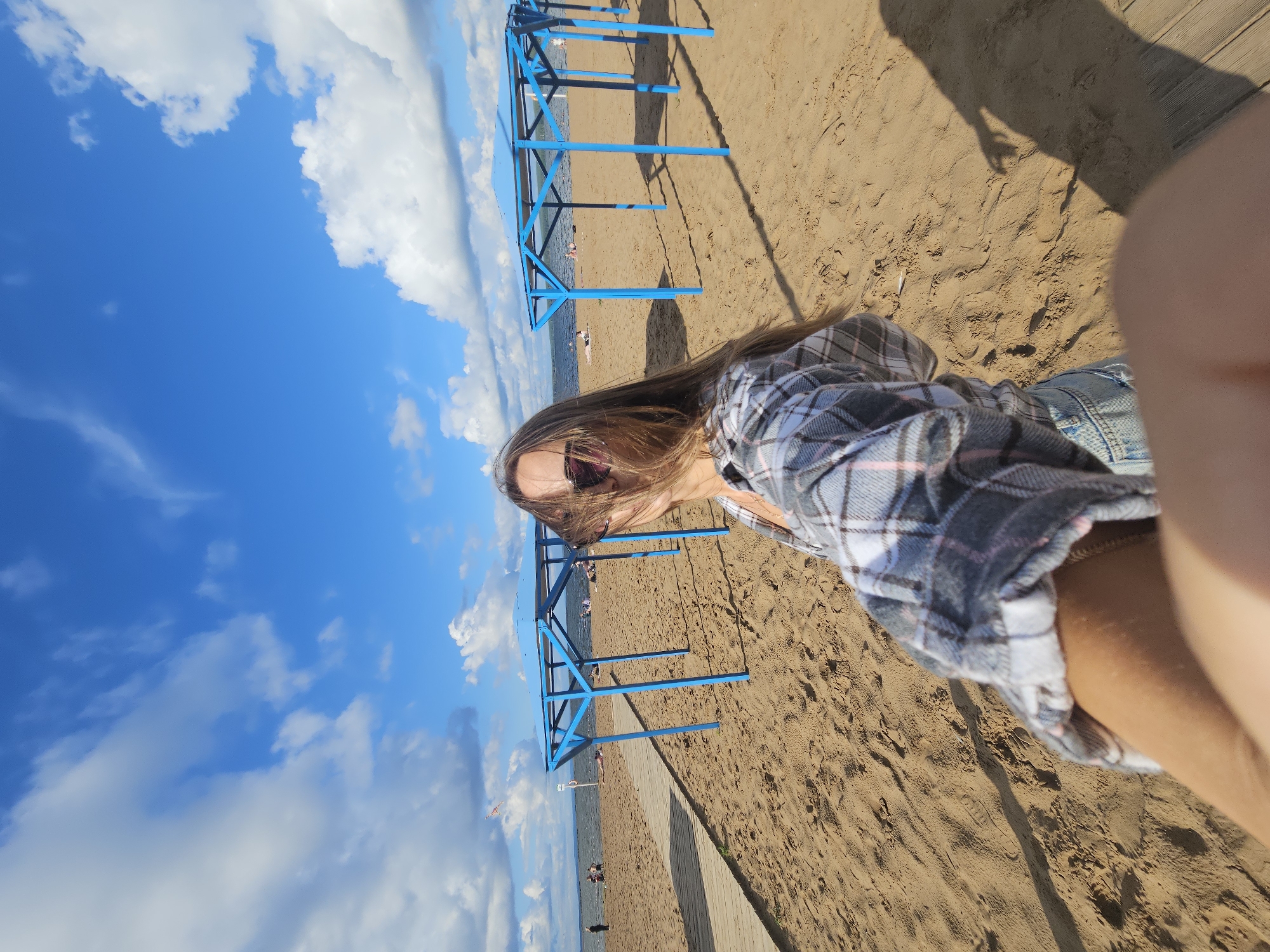 The image depicts a beach scene with a woman in the foreground, holding the camera, taking a selfie. She has long, brown hair and is wearing sunglasses and a plaid shirt. Behind her, the sandy beach stretches out towards a body of water, possibly a river or lake. Several blue beach canopies are visible on the sand, providing shade. In the distance, people are scattered on the beach and in the water. The sky is blue with scattered white clouds. The overall scene suggests a sunny day at a recreational area. The context provided indicates this location is the beach in Chaikovsky, Russia.
