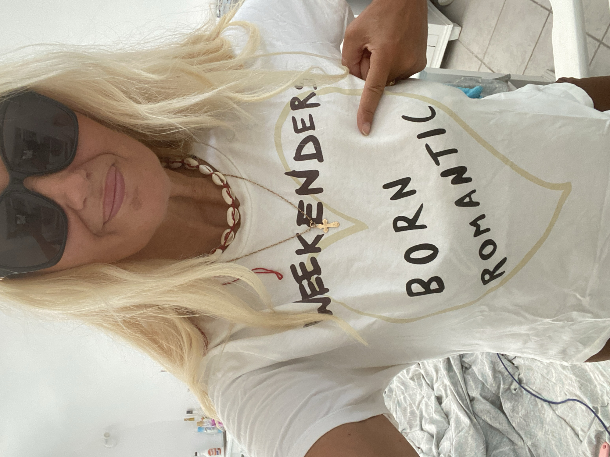 A woman with blonde hair and sunglasses stands indoors, facing the camera and wearing a white t-shirt with the text "WEEKENDERS BORN ROMANTIC" printed on it inside a heart outline. Her right hand is pointing to the text on her shirt. She is wearing a necklace made of seashells. The background appears to be a bedroom with a bed with crumpled linens and a white dresser.