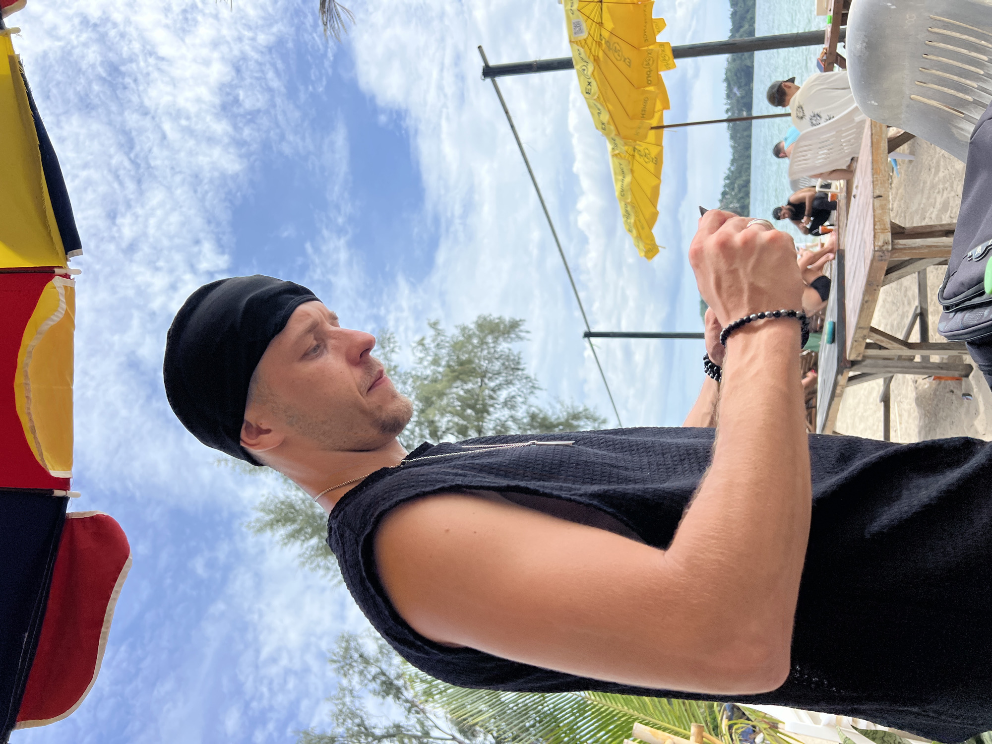 A man wearing a black bandana and a black sleeveless shirt stands on a beach and looks down at his phone, which he holds in his hands. He wears a silver necklace and beaded bracelets on his left wrist. In the background, a wooden table and chairs are set up on the sand, with yellow umbrellas overhead. People are seated at tables in the background, and a body of water is visible. The sky is cloudy with patches of blue.