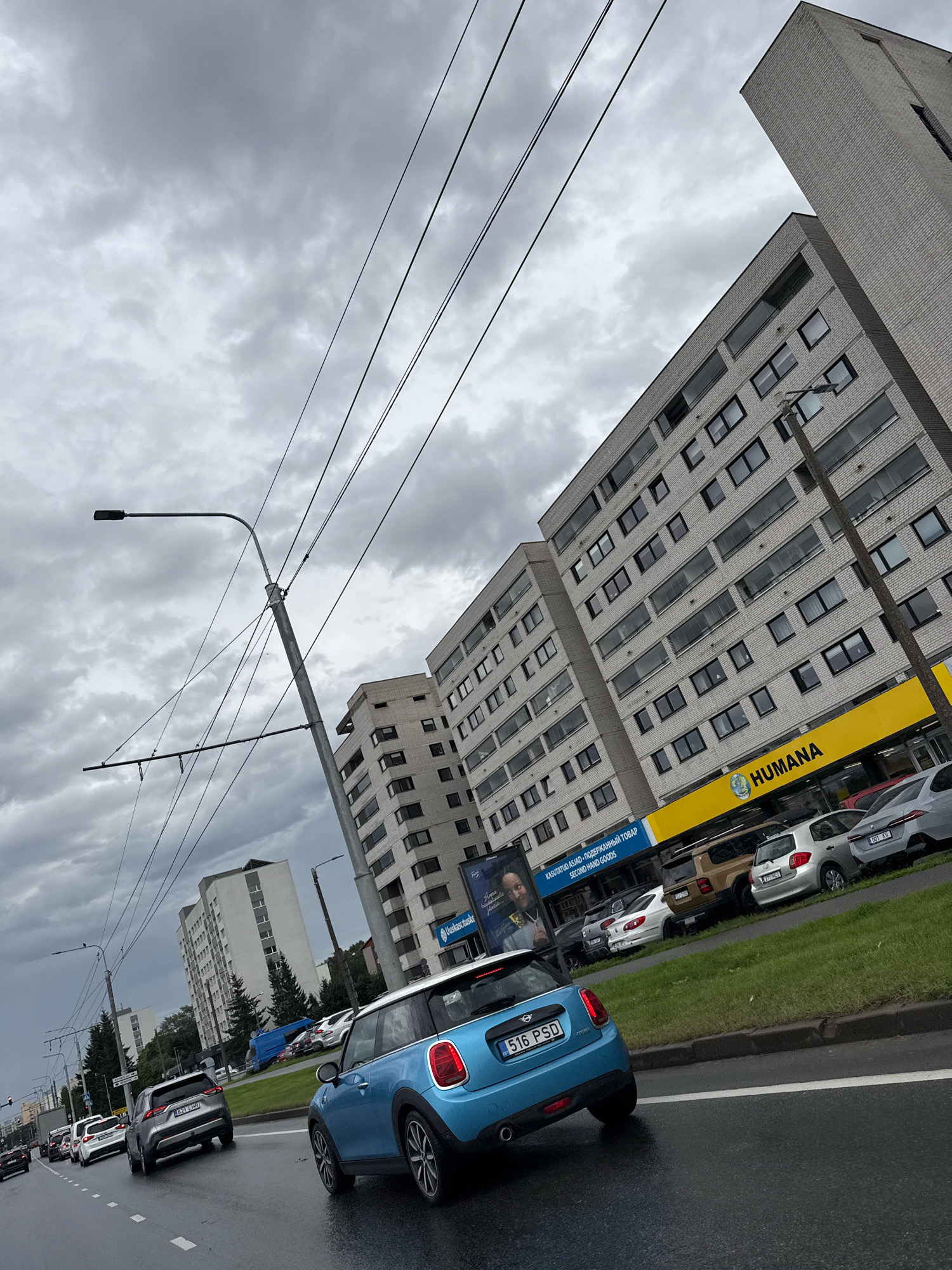 The image depicts a cloudy day in Tallinn, Estonia, with a street view featuring several apartment buildings and cars. The perspective is from a vehicle moving on a wet asphalt road, likely a multi-lane street. Overhead, utility wires stretch across the sky, supported by lampposts.
In the foreground, a blue Mini Cooper is visible, its rear facing the camera. The car's license plate reads "516 PSD". To its left, a grey SUV with license plate "651 LHV" is also on the road. Further back, a line of cars, including white sedans and SUVs, are proceeding along the street.
Dominating the background are large, multi-story apartment buildings constructed with light-colored bricks. One building has a prominent yellow sign that reads "HUMANA," indicating a store. Below this sign, several cars are parked, including a silver sedan with license plate "911 MLF" and a grey SUV. Another building in the background has a sign that says "Uusastimiskeskus".
The overall environment suggests an urban setting with overcast weather. The presence of the utility wires and the style of the apartment buildings are characteristic of residential areas in Eastern Europe. The wet road indicates recent rain.