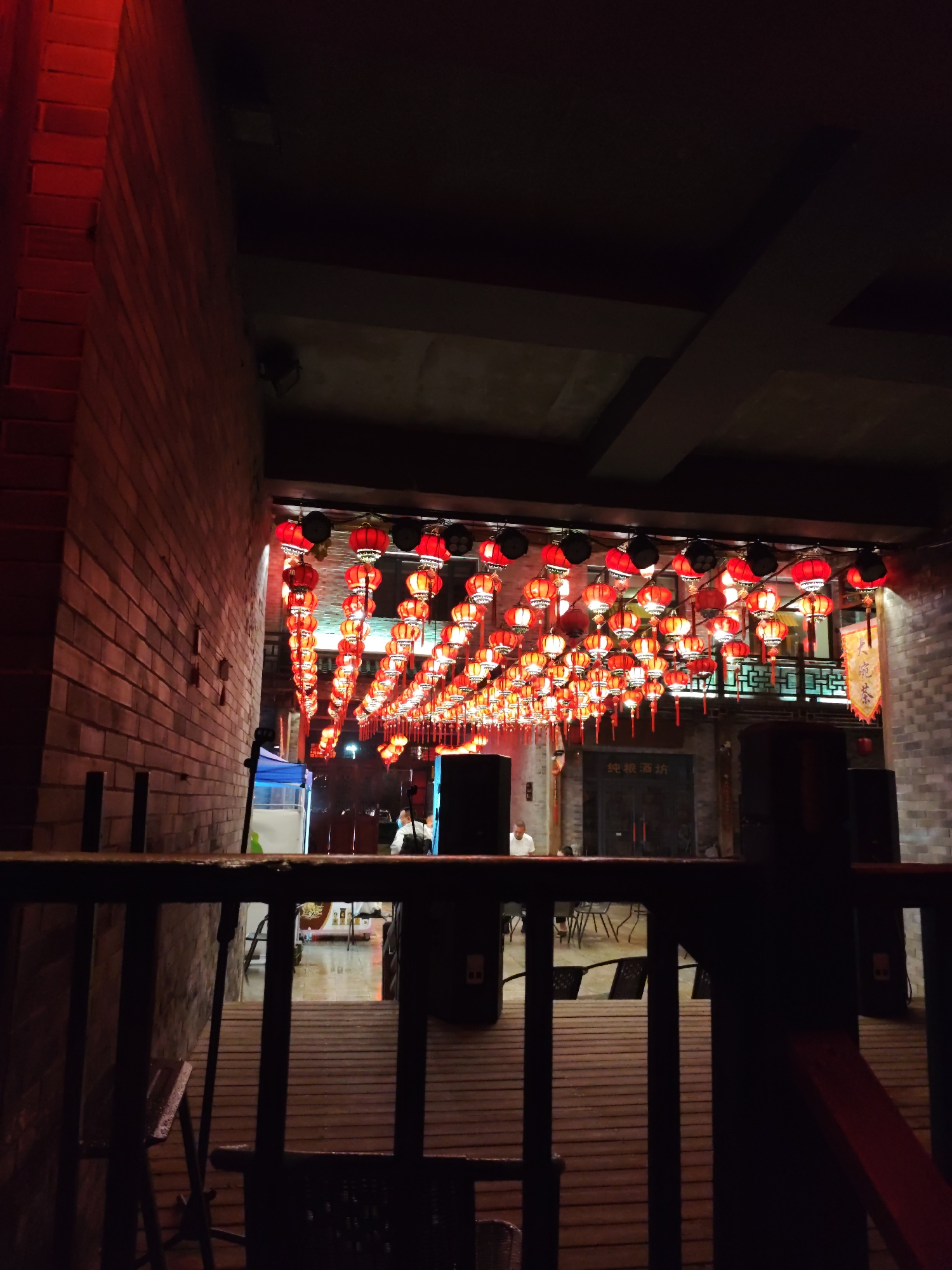 An exterior shot captures a dimly lit street or alleyway in Lin Dian Town, China, adorned with numerous red Chinese lanterns suspended overhead. The lanterns are arranged in rows, creating a festive and illuminated canopy. To the left, a brick wall, illuminated by a red light, borders the scene. In the background, a building with traditional Chinese architectural elements is visible, featuring dark wooden accents and intricate railings. Several figures are present, appearing to be patrons or staff, engaged in various activities, though their actions are not clearly discernible due to the low light. The ground appears to be paved or made of wooden planks, with some furniture, including tables and chairs, set up in the open space. The overall atmosphere suggests a nighttime scene in a commercial or entertainment district.