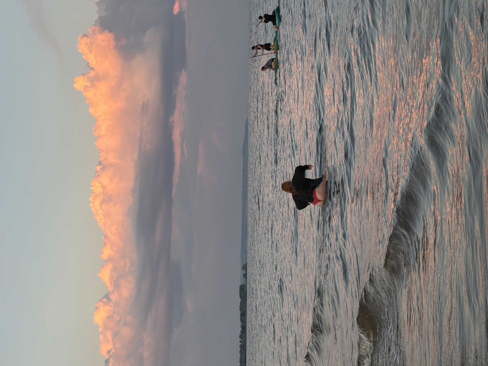 In Laishevo, Russia, at sunset, several people are seen engaging in water activities. A person wearing a black top and red bottoms is in the foreground, waist-deep in the water, facing away from the camera. In the mid-ground, three individuals are on paddleboards, with two standing and one kneeling, paddling across the calm, rippling water. The horizon is lined with trees and a distant landmass. The sky above is a gradient of soft blues and pinks, illuminated by the setting sun, with dramatic, billowy clouds catching the warm light. The water reflects the colors of the sky, creating a serene and picturesque scene.