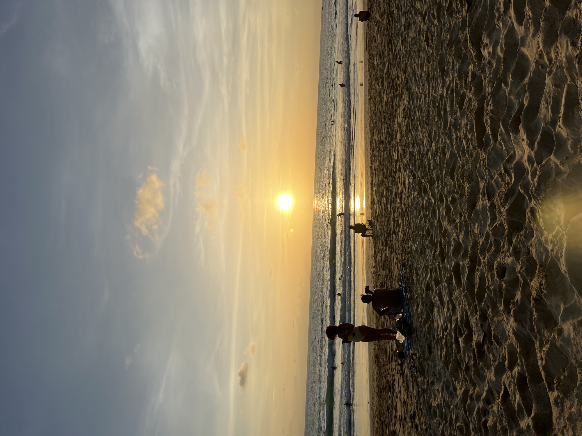 A silhouette of a woman in a bikini stands on a sandy beach in Choeng Thale, Thailand, facing the ocean during sunset. A man sits on the sand behind her, looking at his phone. The sun is low in the sky, casting a golden glow on the water and clouds. Waves are gently rolling in, and several people are visible in the water, some surfing. The sand is textured with footprints and ripples.