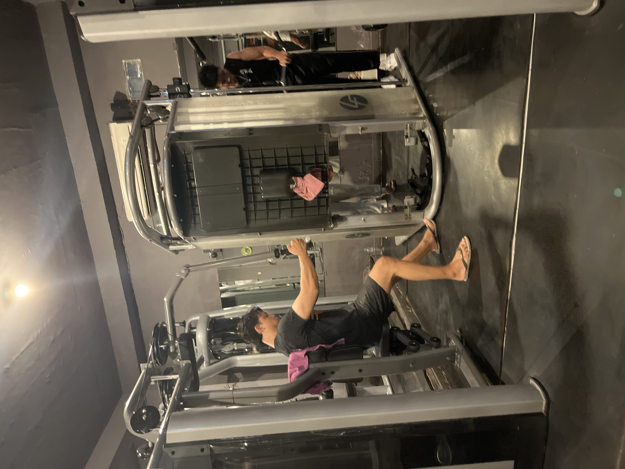 A man in a black t-shirt and shorts is seated on a weight machine, holding the handles and performing a chest exercise. A pink towel is draped over the back of the seat. Another man in a black tank top and pants is standing and operating a weight machine nearby, in a gym. The gym is dimly lit with a bright light fixture visible on the ceiling. The floor is black and the gym equipment is silver. The scene appears to be in a gym located in Denpasar, Indonesia.