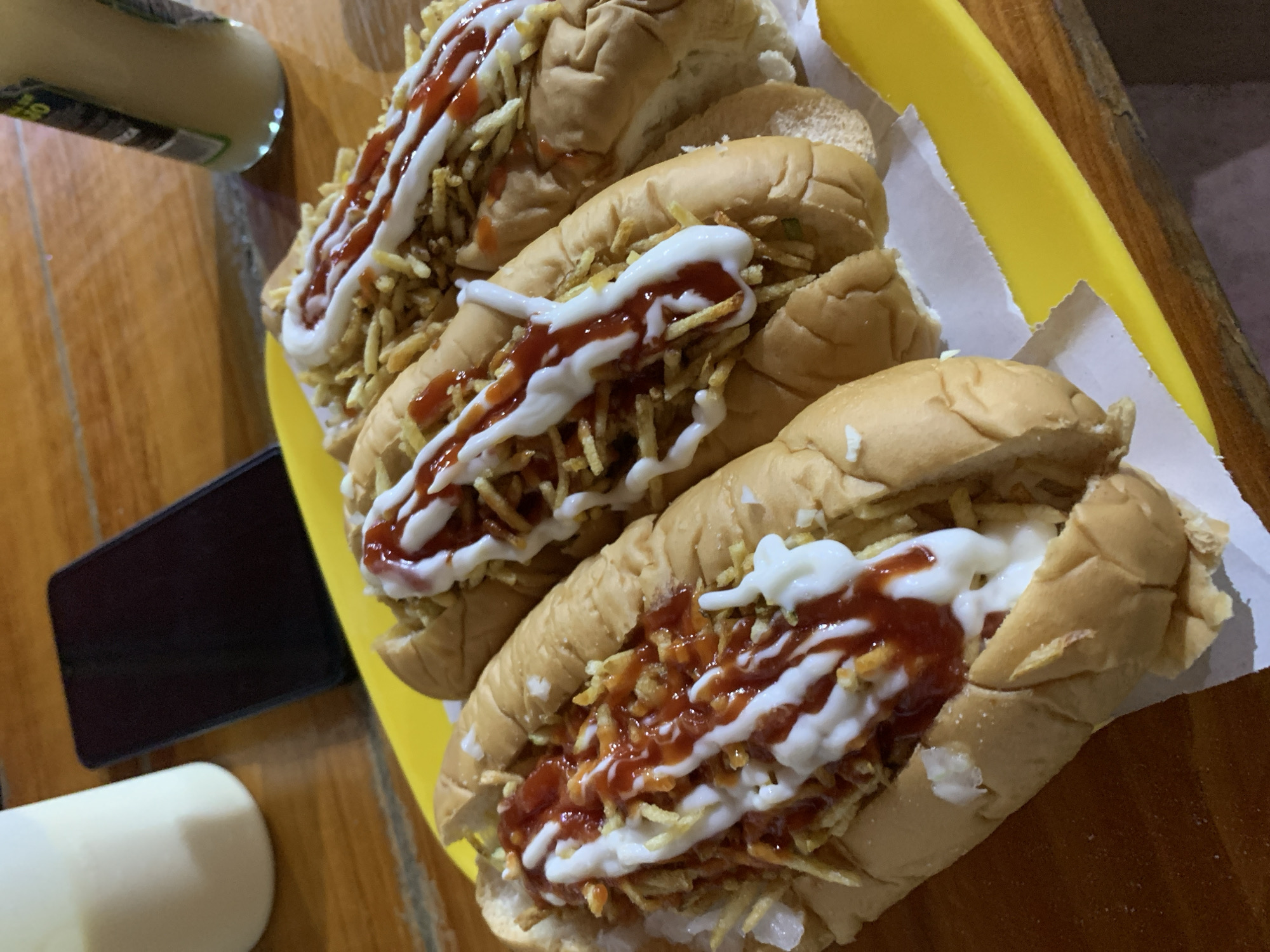 An overhead shot depicts four generously topped hot dogs arranged on a yellow tray lined with white paper. Each hot dog is served in a soft, elongated bun and filled with a sausage, then covered with a layer of thin, crispy potato sticks. Drizzled over the potato sticks are contrasting zigzag patterns of bright red ketchup and white mayonnaise.
The tray rests on a wooden table, which appears to be a light brown color. In the upper left corner of the frame, a white bottle, possibly containing a sauce or beverage, is partially visible. Further down on the left side, a black smartphone lies flat on the table, next to a portion of a white mug or cup.
The lighting in the image is somewhat dim, suggesting an indoor setting, possibly during the evening or night, creating a casual atmosphere. The style of hot dogs, with crispy potato sticks and multiple sauces, is characteristic of street food often found in Santa Ana, Venezuela, matching the provided location context. No legible text is visible on any objects. The scene captures a moment before or during a casual meal.