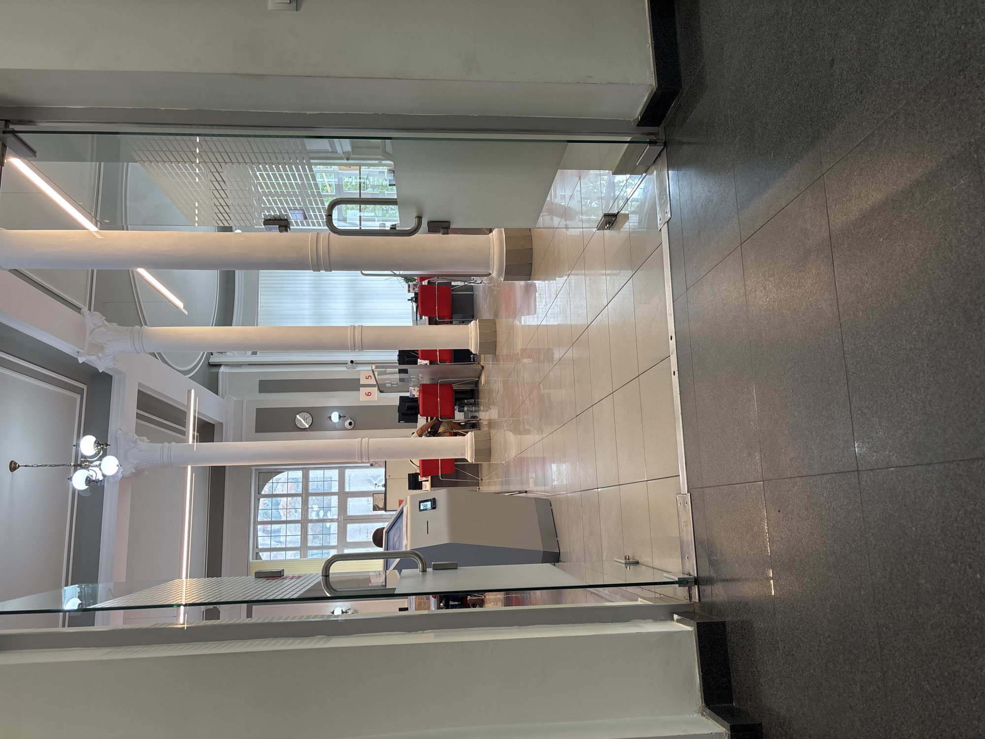 The image shows the interior of a modern public building, likely a bank or administrative office, with a view through a glass door. The interior features white tiled floors and large white columns, typical of neoclassical architecture. In the background, several workstations with red chairs are visible, suggesting a service or administrative area. A self-service kiosk stands near the entrance. The ceiling has decorative molding and modern lighting fixtures. The overall impression is one of a well-maintained, public space. The location of this building in Pleven, Bulgaria, is not evident from the image itself.