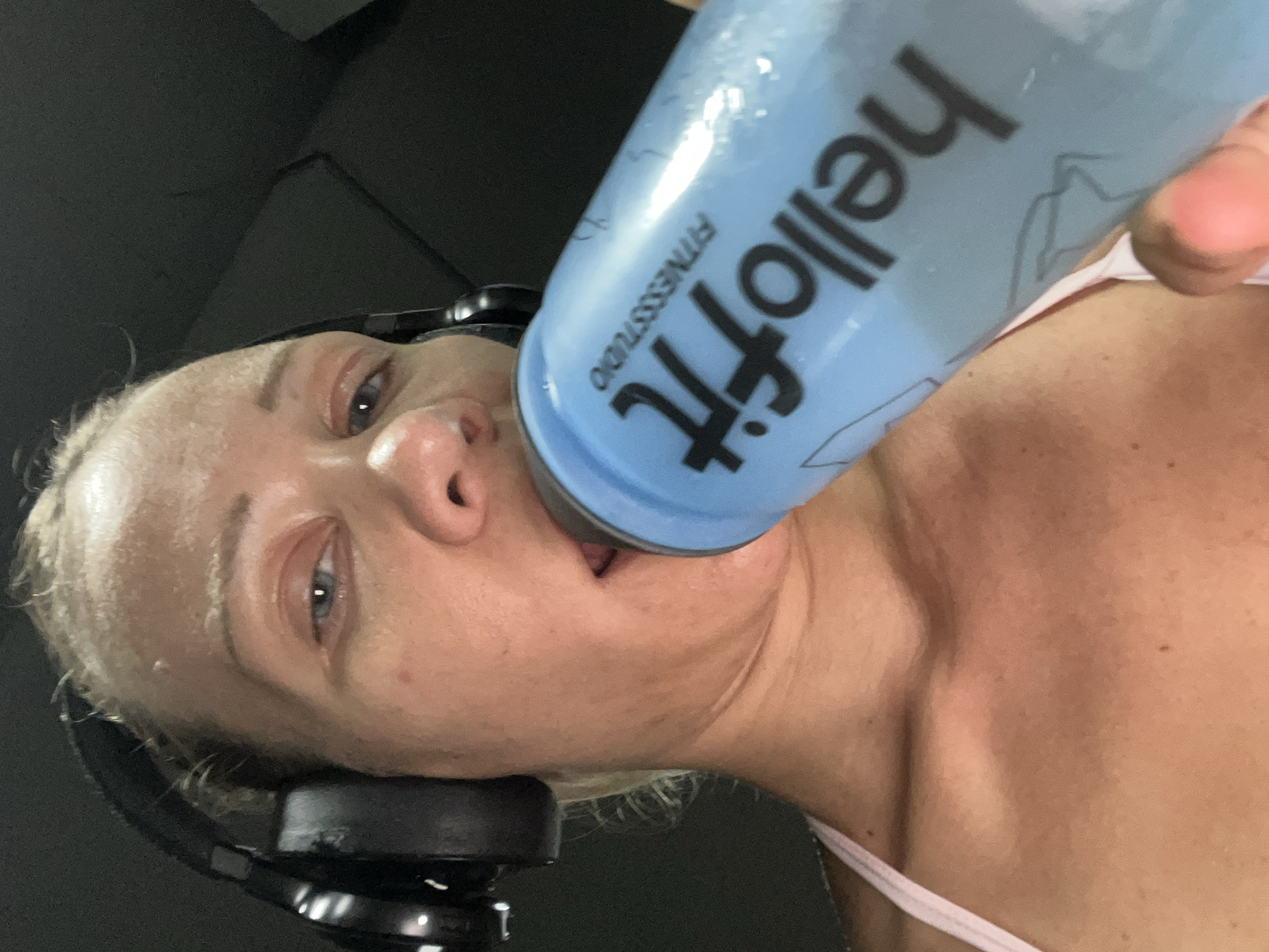 A selfie-style, close-up shot shows a fair-skinned woman with blonde hair, wearing black headphones, and holding a light blue water bottle to her mouth. The bottle has black text that reads "hellofit FITNESSSTUDIO". She appears to be in an indoor environment, possibly a gym, with dark, indistinct backgrounds. The image is lit from above and to the left, creating highlights on her forehead and the water bottle. The woman is wearing a light pink athletic top. There are no identifiable landmarks or contextual clues within the image that would indicate a specific location in Altötting, Germany.