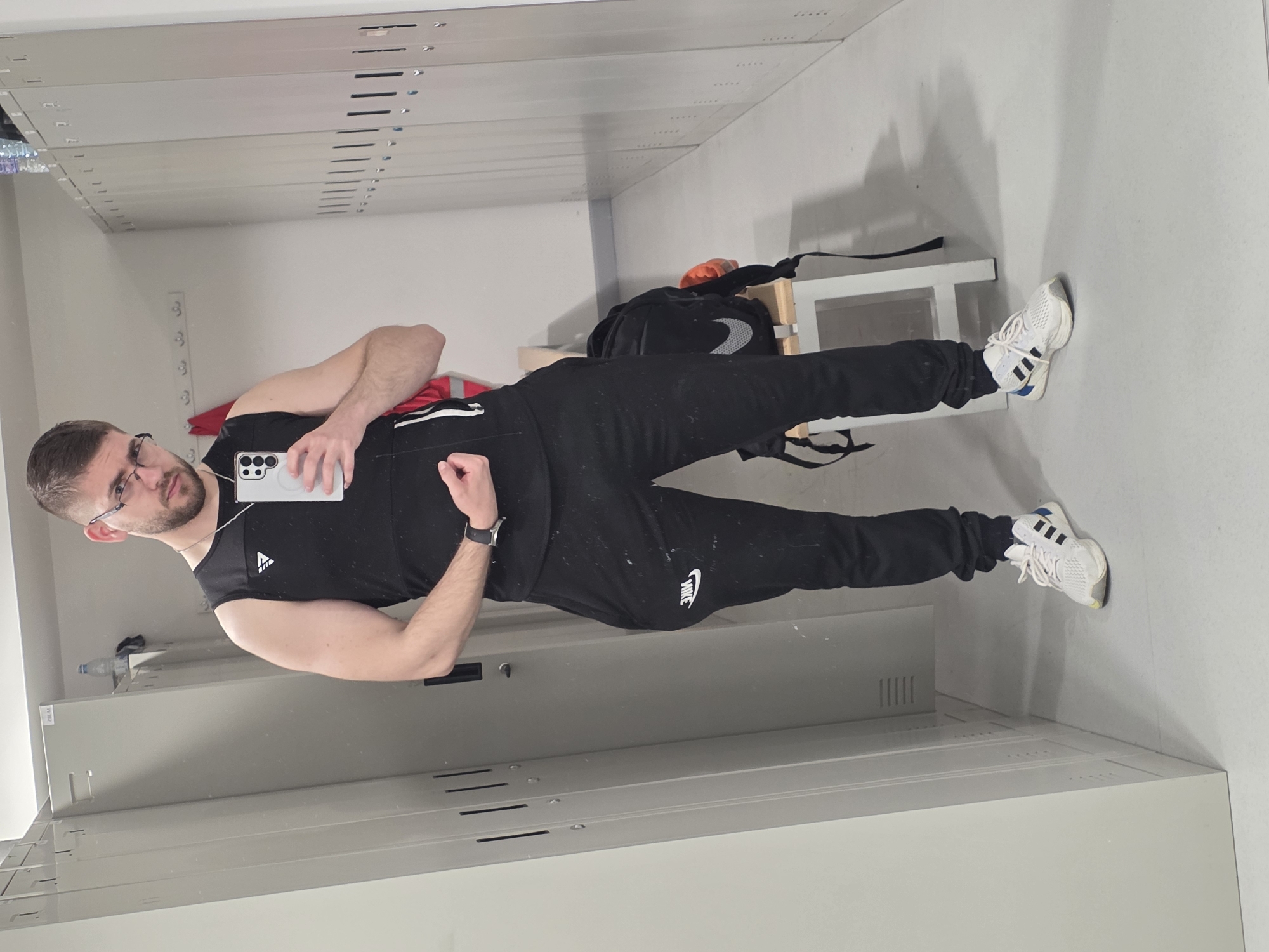 A young Caucasian man is featured in a mirror selfie, taken in an indoor setting resembling a locker room or gym facility in Plovdiv, Bulgaria. The image is oriented sideways, with the man standing upright, looking directly into the camera of his smartphone.
He has short dark hair, a neatly trimmed beard, and wears clear-framed glasses. He is dressed in athletic attire: a black sleeveless top with a sports brand logo (resembling Adidas) on the chest, and black track pants with the white "NIKE" logo prominently displayed on the left thigh. On his left wrist, he wears a dark smartwatch. His feet are clad in white athletic shoes with black stripes and blue accents, characteristic of Adidas footwear. He is holding a light-colored Samsung Galaxy smartphone with multiple camera lenses in his right hand.
The background shows a brightly lit room with light-colored walls and floor. Rows of light grey metal lockers or storage units line the walls, some featuring vents or horizontal slats. Behind the man, a white bench or shelf holds a black backpack with a white Nike swoosh and a red strap or item. A white water bottle is partially visible on a shelf to the left. The man's expression is neutral and direct, suggesting he is posing for the photo.
