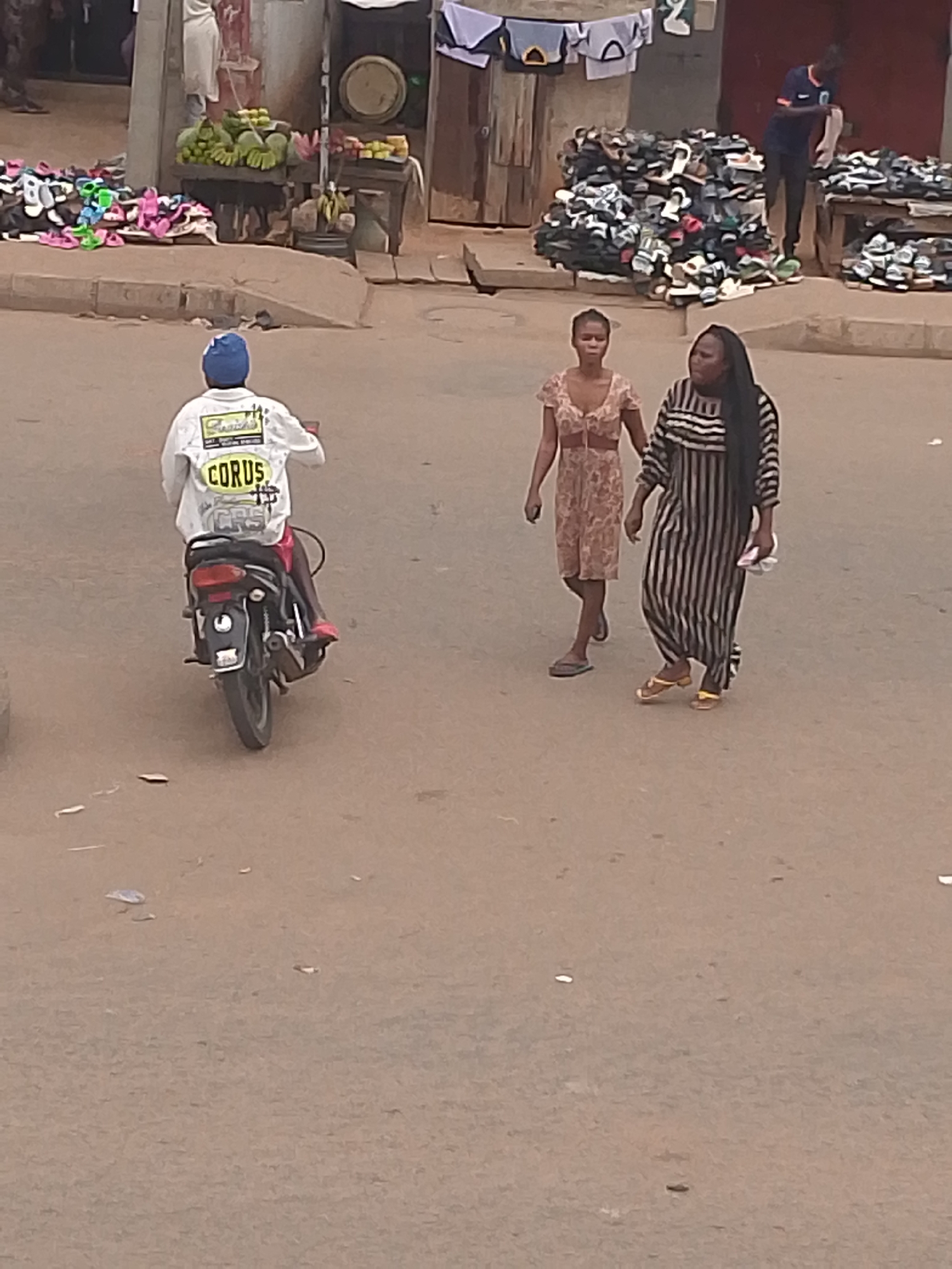 The image depicts a street scene in Bukuru, Nigeria. The ground is a dusty brown color. A person on a motorcycle is moving away from the camera. The person is wearing a blue hat and a white jacket with the word "CORUS" on the back. Two women are walking toward the right of the image. One woman wears a brown patterned dress, and the other wears a long, vertically striped black and white dress. Street vendors with goods for sale are visible in the background, including piles of sandals and fruit. A man in a blue shirt is near the sandals vendor.