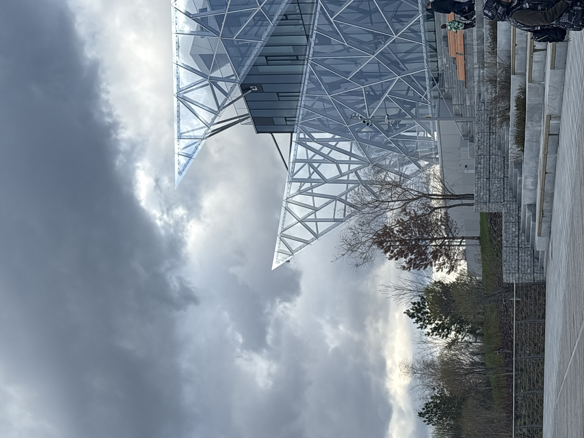 The image shows a modern building with a glass and steel frame in Kazan, Russia. The building is characterized by a complex, geometric design with triangular patterns in its facade. The sky is overcast with heavy, gray clouds. Near the building, there are trees with bare branches. A staircase with stone steps leads up to the building. There are indistinct figures walking along the top of the staircase. A pathway or sidewalk is visible in the lower right of the frame.