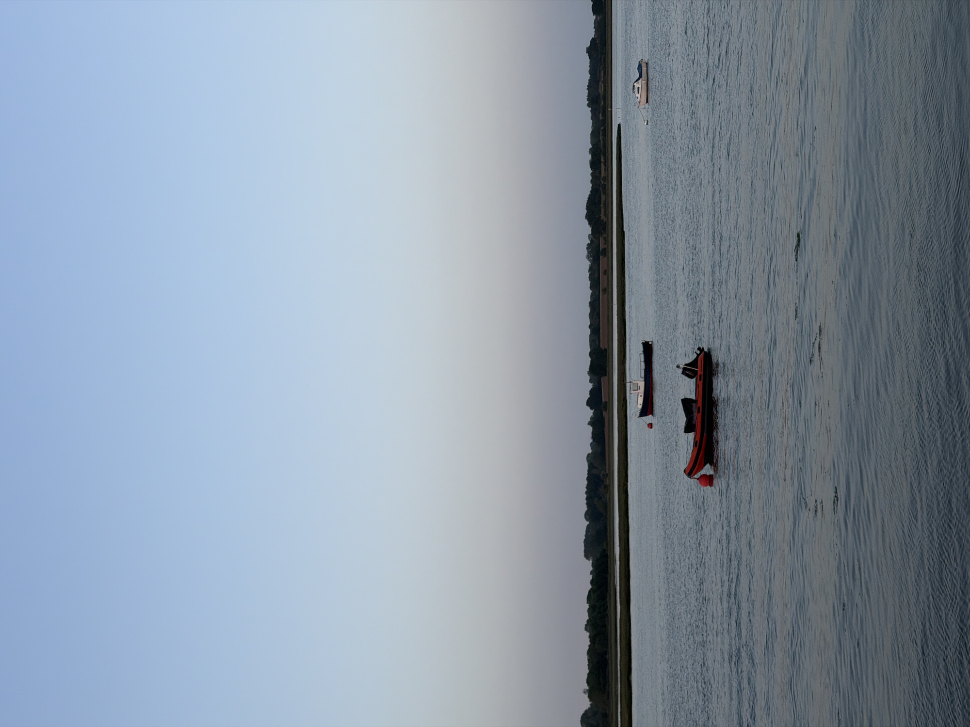 The image depicts a waterway in East Suffolk, United Kingdom, with a sliver of land running vertically through the middle of the frame. The left side of the image shows a sky gradient from light blue at the top to a light grey near the land. The right side shows water with small waves. Three boats are visible on the water: a larger, light-colored boat towards the top, a smaller white and red boat near the middle, and an orange and black boat near the bottom. The land is lined with trees.