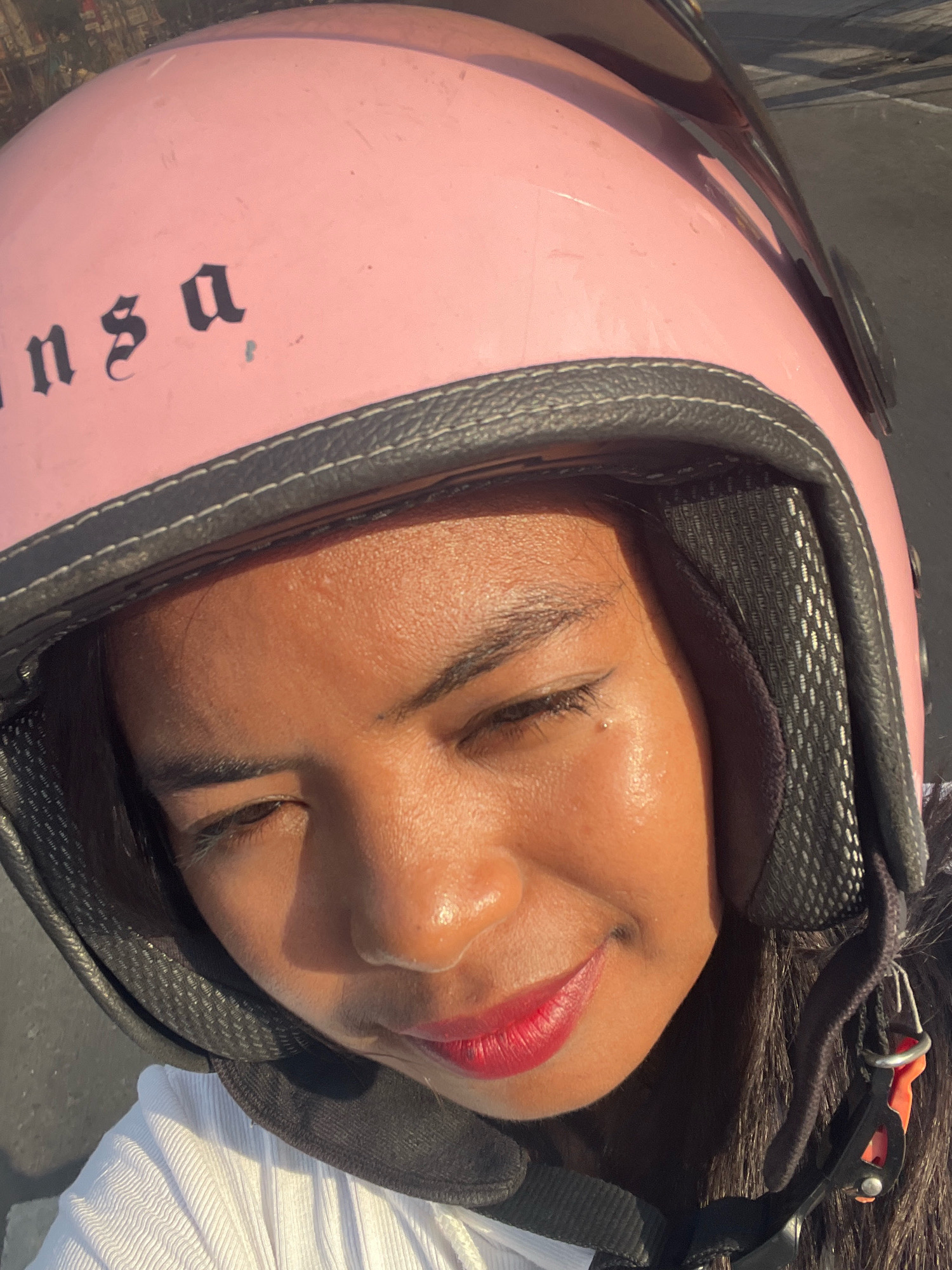 A medium shot captures a person wearing a pink motorcycle helmet in Kuta, Indonesia. The helmet has black lettering that reads "ansa" in a gothic font. The person's face is visible, with their eyes closed, and they are wearing red lipstick. The image is taken outdoors, with sunlight casting shadows. The background suggests an urban environment, possibly a street or pavement.