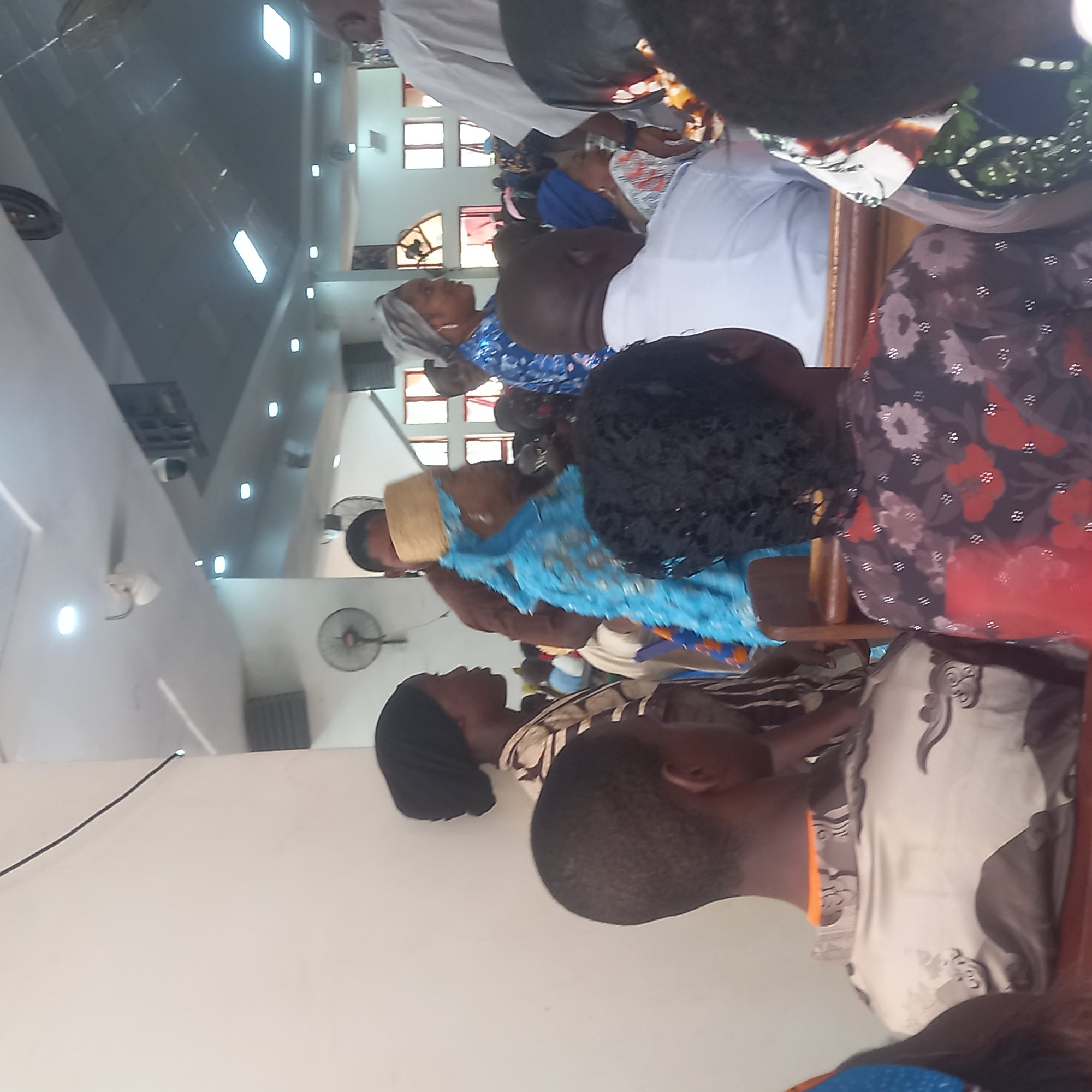 A gathering of people is indoors, likely in Barden Yana, Nigeria. The setting appears to be a church or a similar religious space, indicated by the rows of seating and interior architecture. Individuals are dressed in a variety of clothing styles, some wearing traditional Nigerian attire. The room is brightly lit, with artificial lighting supplemented by daylight filtering through windows. Ceiling fans are present. The people are sitting attentively, possibly participating in a service or event. Some are facing forward, focused on something outside the frame. The environment is clean and well-maintained.