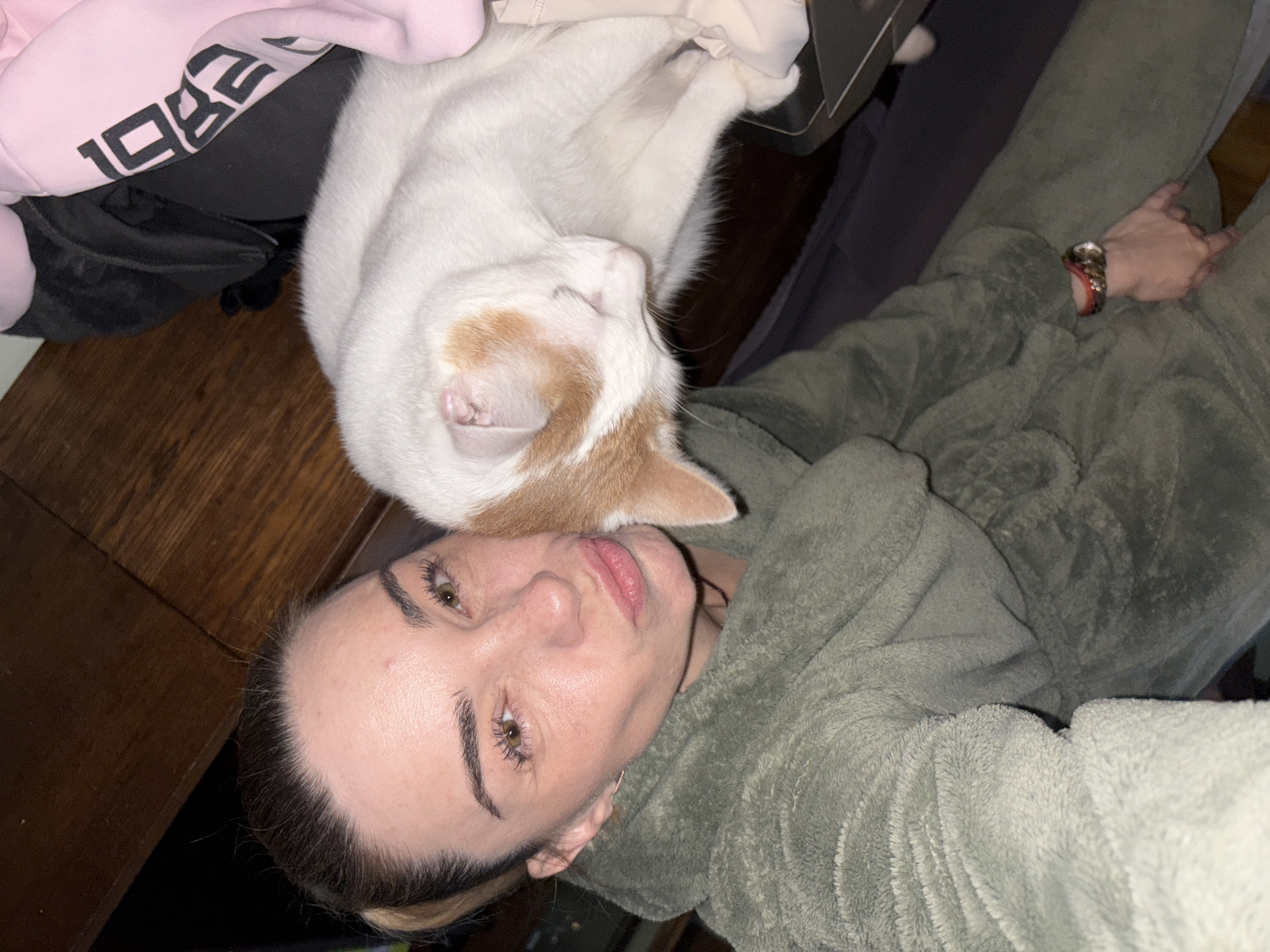 In an indoor, dimly lit environment, a woman with her hair pulled back in a ponytail and wearing a green bathrobe is taking a selfie. She is making a kissing face towards the camera. A white and orange cat is nuzzled against her cheek, its eyes closed. Behind the woman, a pink hoodie with the text "1982" printed in black is visible, along with dark clothing and wooden furniture. The overall scene suggests a relaxed, casual moment at home.