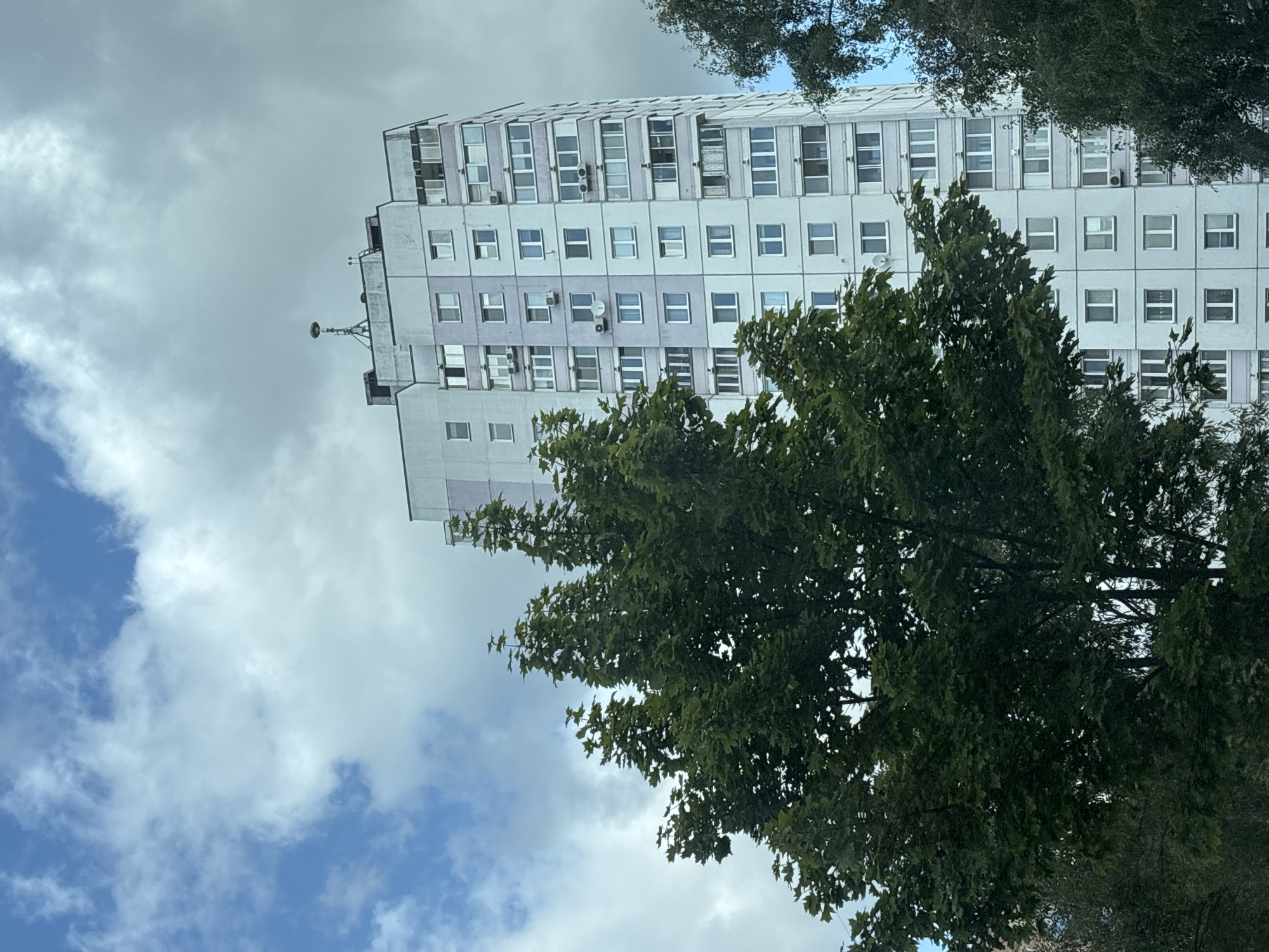 A tall, light purple and white apartment building stands partially obscured by a large, dark green tree in the foreground. The building has many windows and balconies. On top of the building, there is a tall antenna with a light fixture. The sky above is filled with grey and white clouds, with patches of blue visible. The scene appears to be located in an urban or suburban environment.