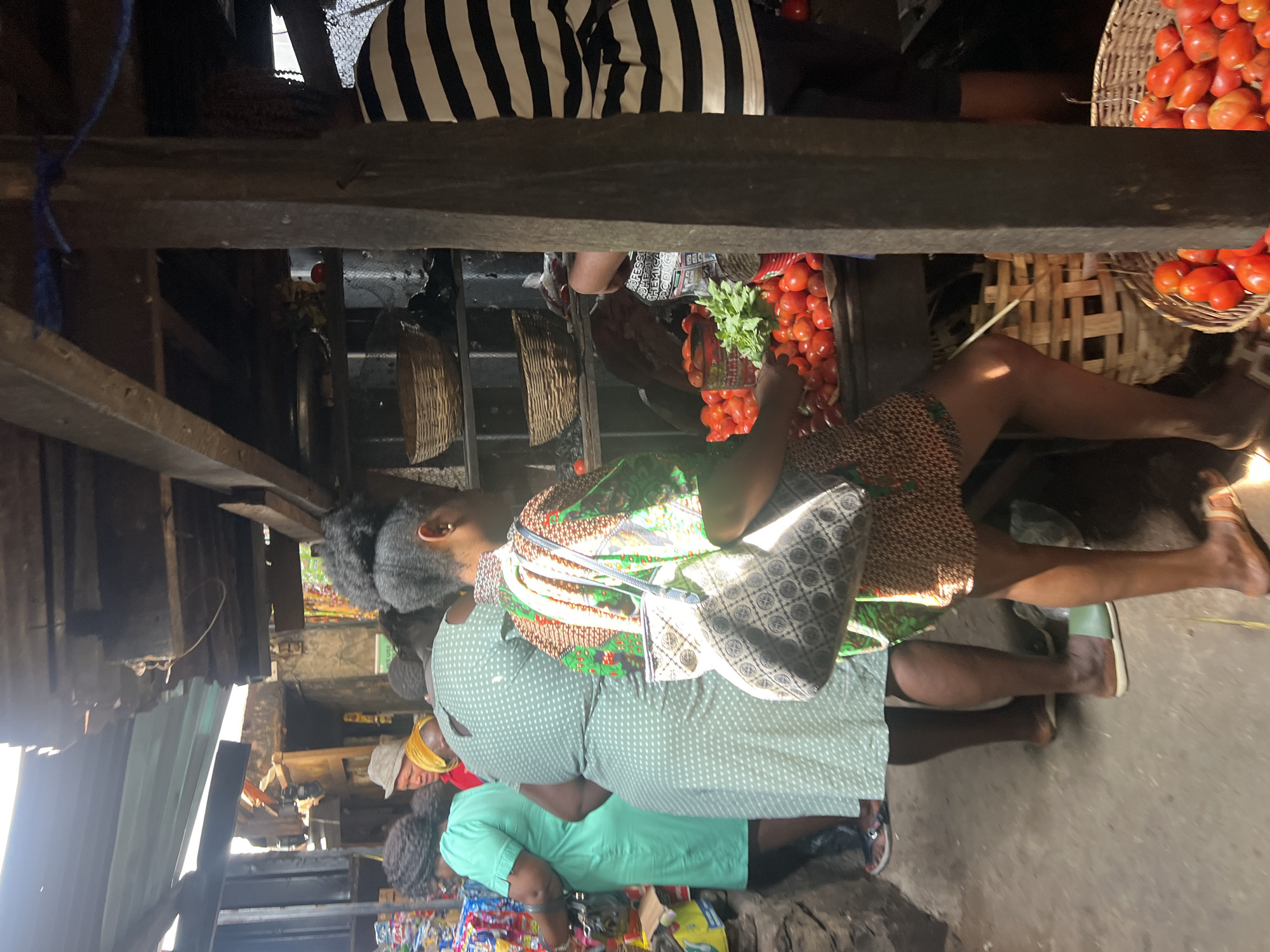 In Nkpor, Nigeria, a market scene unfolds under a makeshift, partially dilapidated wooden and corrugated metal roof. Sunlight streams through gaps, illuminating dust motes and casting shadows on the dirt ground. Several people are present. In the foreground, a woman in a patterned dress with a bag slung over her shoulder examines a pile of red tomatoes at a stall. Behind her, another woman in a green polka-dot dress stands beside a stall filled with goods, and further to the left, a woman in a bright green dress is also browsing. Men and other figures are visible in the background, including one wearing a hat and another in a striped shirt. The market stall features woven baskets, stacked goods, and fresh produce, predominantly tomatoes. The overall impression is of a bustling, everyday market environment.