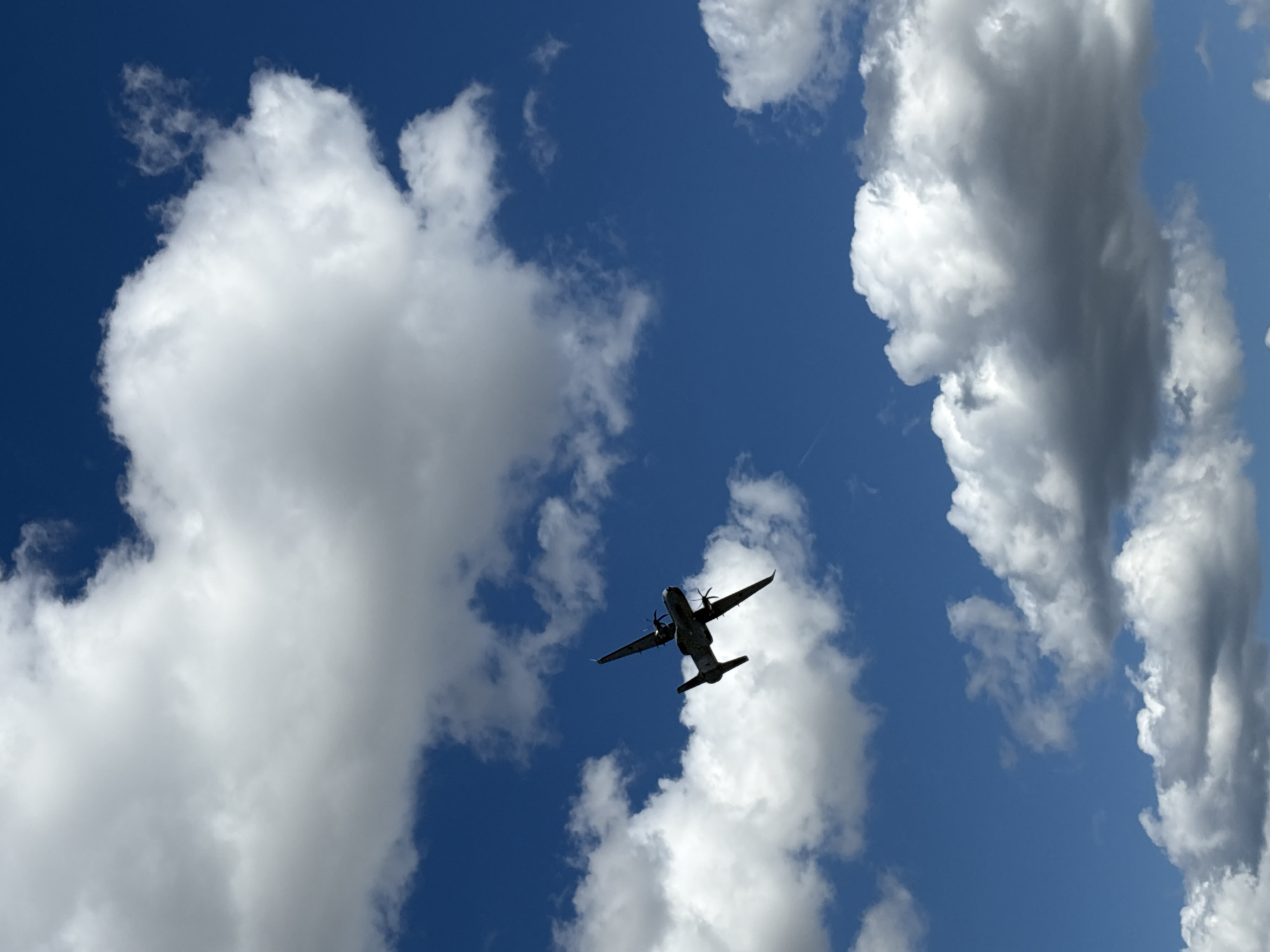 An airplane flies through a blue sky dotted with white, fluffy clouds. The airplane, silhouetted against the clouds, is a twin-engine propeller aircraft. It appears to be in flight, possibly performing aerobatics given its tilted position. The image was taken on a sunny day, as indicated by the bright blue sky and the well-defined clouds. The exact location in Cheb, Czechia cannot be determined from the image alone.
